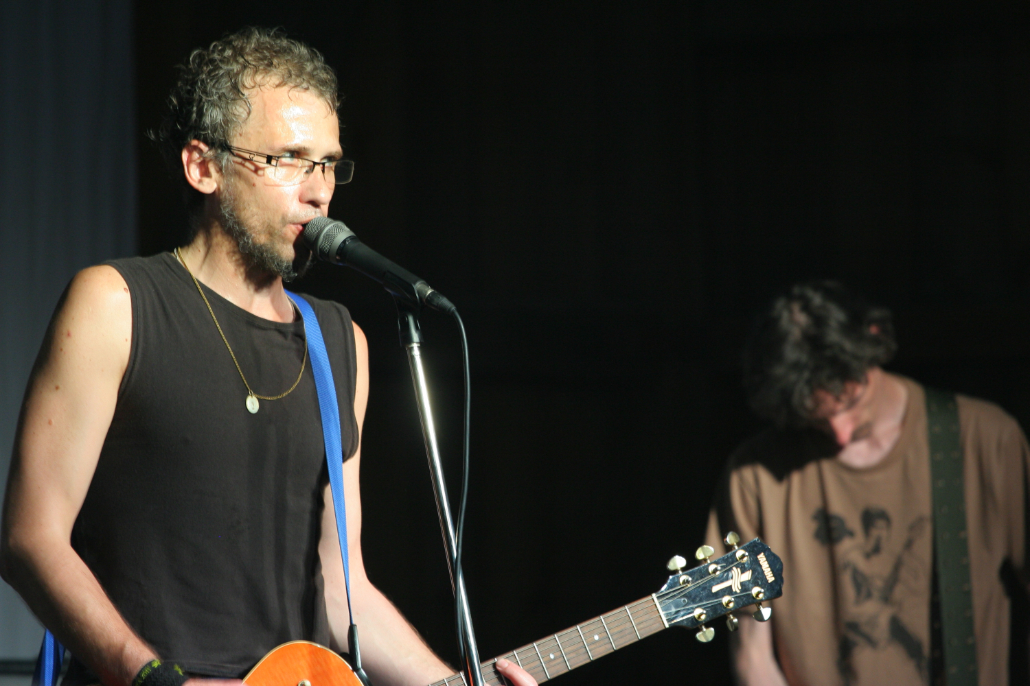 A man with glasses and a beard is playing an electric guitar and singing into a microphone. He is wearing a black sleeveless shirt, a necklace, and jamming out like it's 2011. Another man in the background, in a brown t-shirt, is also playing guitar. The dimly lit setting adds to the Laba daba vibe.