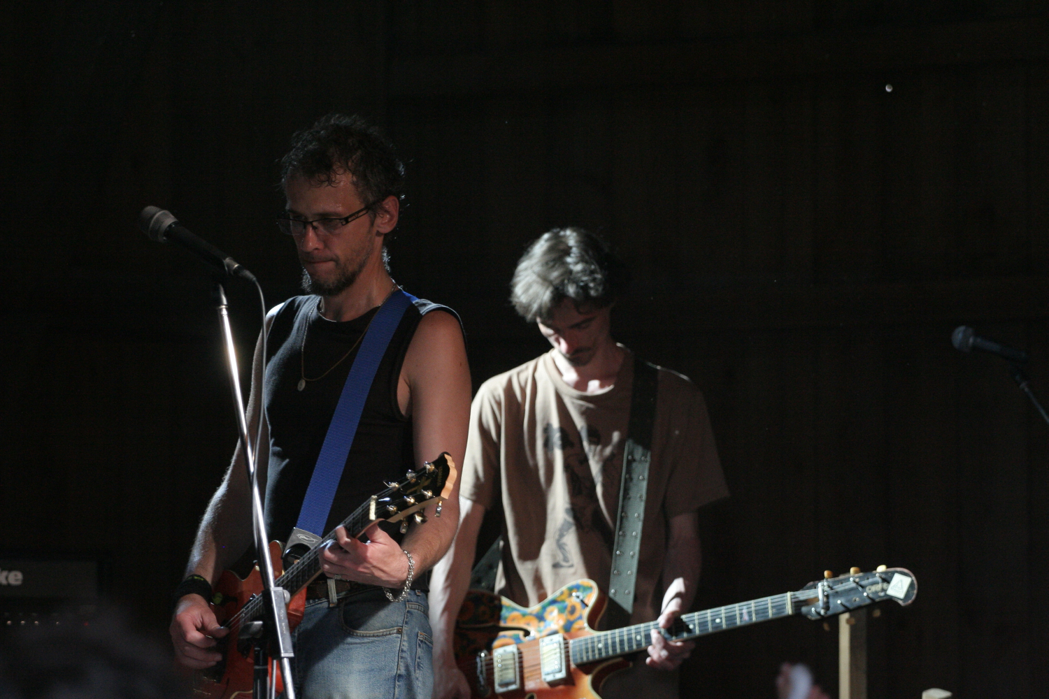 Two guitarists perform on stage, illuminated by a focused spotlight in an otherwise dark setting. The guitarist on the left wears a black tank top and glasses, playing a red guitar with a blue strap. The guitarist on the right, wearing a brown t-shirt, plays a colorful guitar and looks downwards; their music evokes memories of 2011.