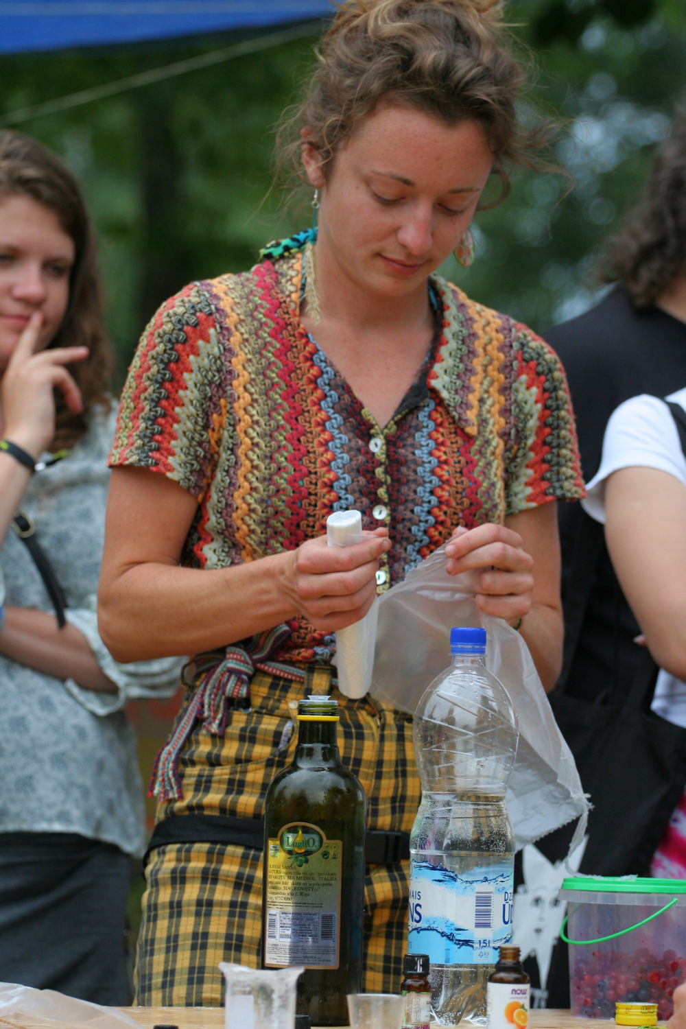 A person with curly hair, wearing a colorful knit top and checkered pants, is using a white plastic tool, possibly a knife or peeler, on a translucent plastic bag at an outdoor Laba event. Bottles and containers are on the table in front, with onlookers in the background.