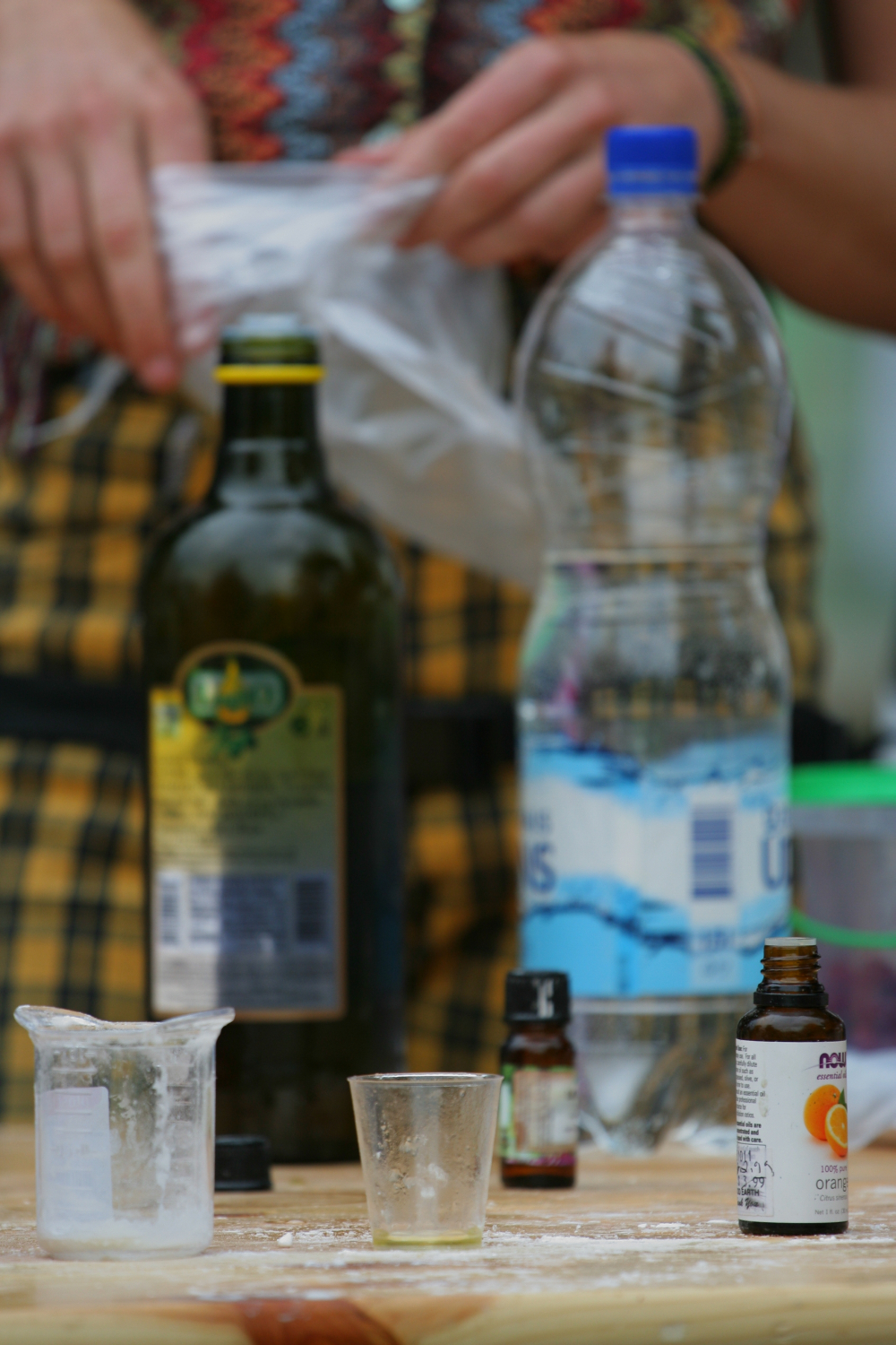 Close-up of a table containing various items: a bottle of olive oil, a plastic bottle of water, a small glass, a measuring cup with white powder, and small bottles of essential oils. Each item is placed on a wooden surface. In the background, someone in a colorful apron manages a Laba Daba labeled bag from 2011.
