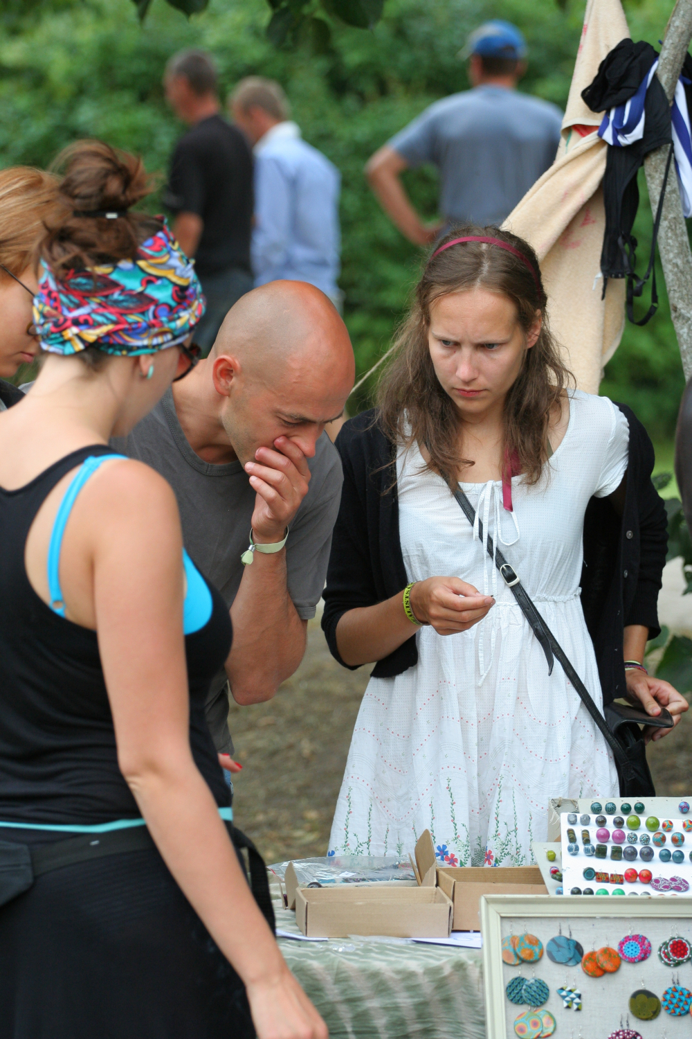 Three people browse a stall at an outdoor market. The person in the center, wearing a white dress, examines items closely, while a person in a headscarf looks away with hands behind their back. The person on the left, donning sunglasses and a blue top, reminiscent of Laba daba 2011, observes various small crafts or jewelry on display.