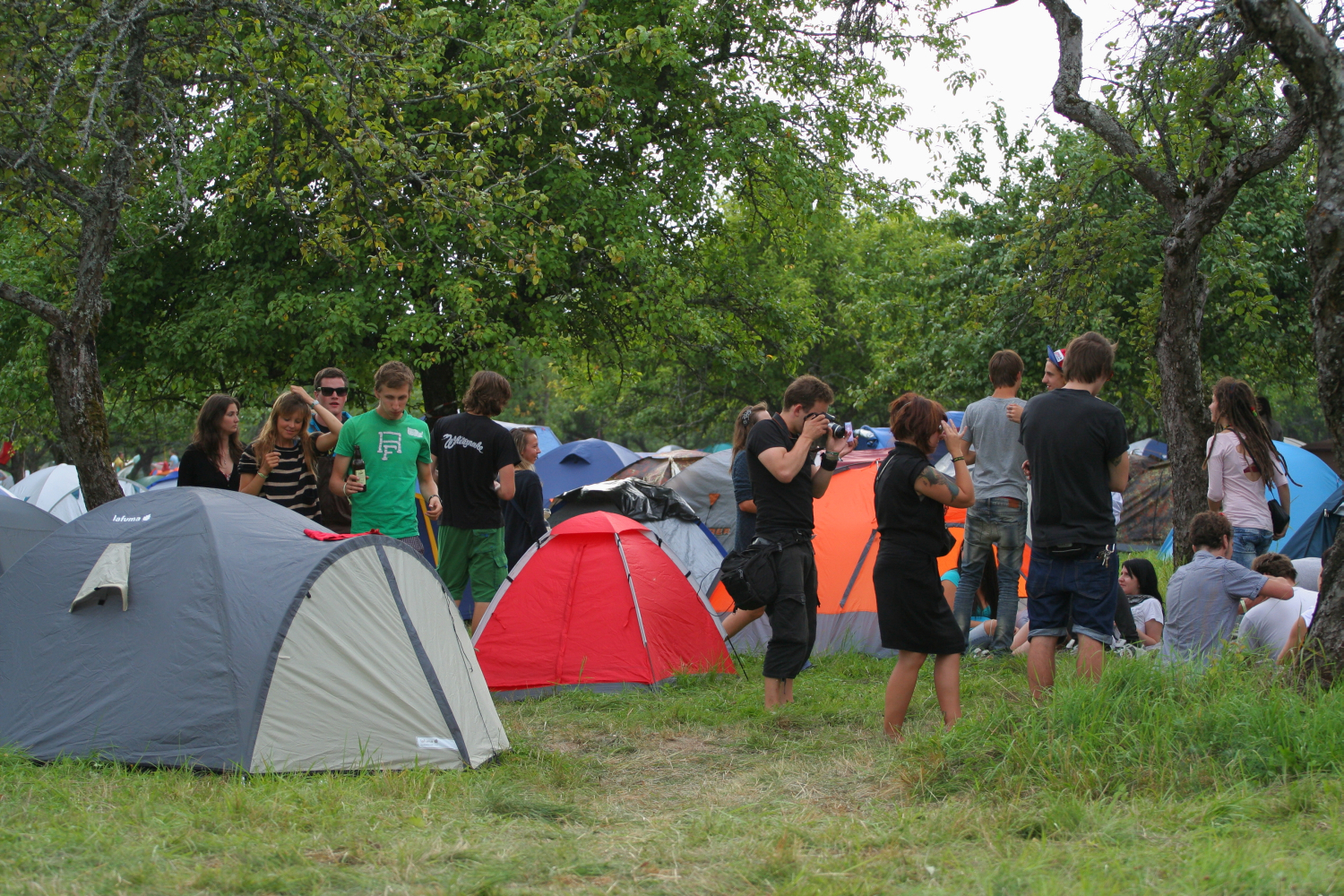 A group of people at an outdoor campsite with several colorful tents, including red, gray, orange, and blue. Laba daba 2011 is in full swing as some people stand and converse while others sit on the grass. Trees surround the area, providing shade and a sense of seclusion. The atmosphere seems relaxed and social.