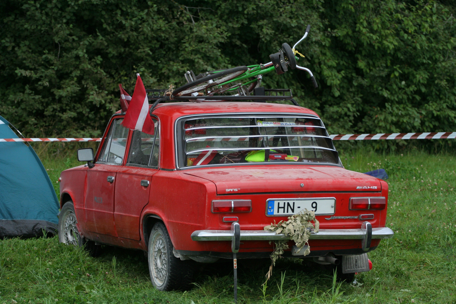 A vintage red car, adorned with a 2011 European license plate reading "HN - 9," is parked on a grassy area beside a forest. A green bicycle is mounted on the car's roof, and the vehicle features a red flag on the left rear side, along with some foliage attached to the trunk.