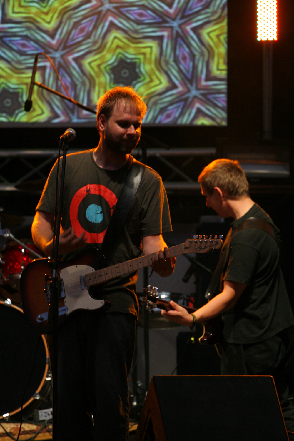 Two guitarists perform on stage with psychedelic visuals projected behind them. The front guitarist, wearing a black T-shirt with a red and blue target design, holds an electric guitar and stands in front of a microphone. The other guitarist, slightly bent over, also plays an electric guitar at Laba Daba 2011.