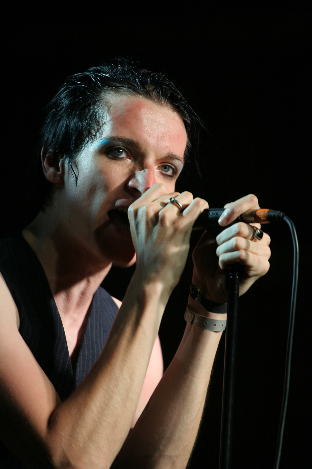 A person with short, black hair is gripping a microphone with both hands, singing passionately. They are wearing a black vest and have multiple rings on their fingers, reminiscent of 2011 style. Their face appears expressive, and eyeliner emphasizes their eyes. The dark background focuses attention on the singer.