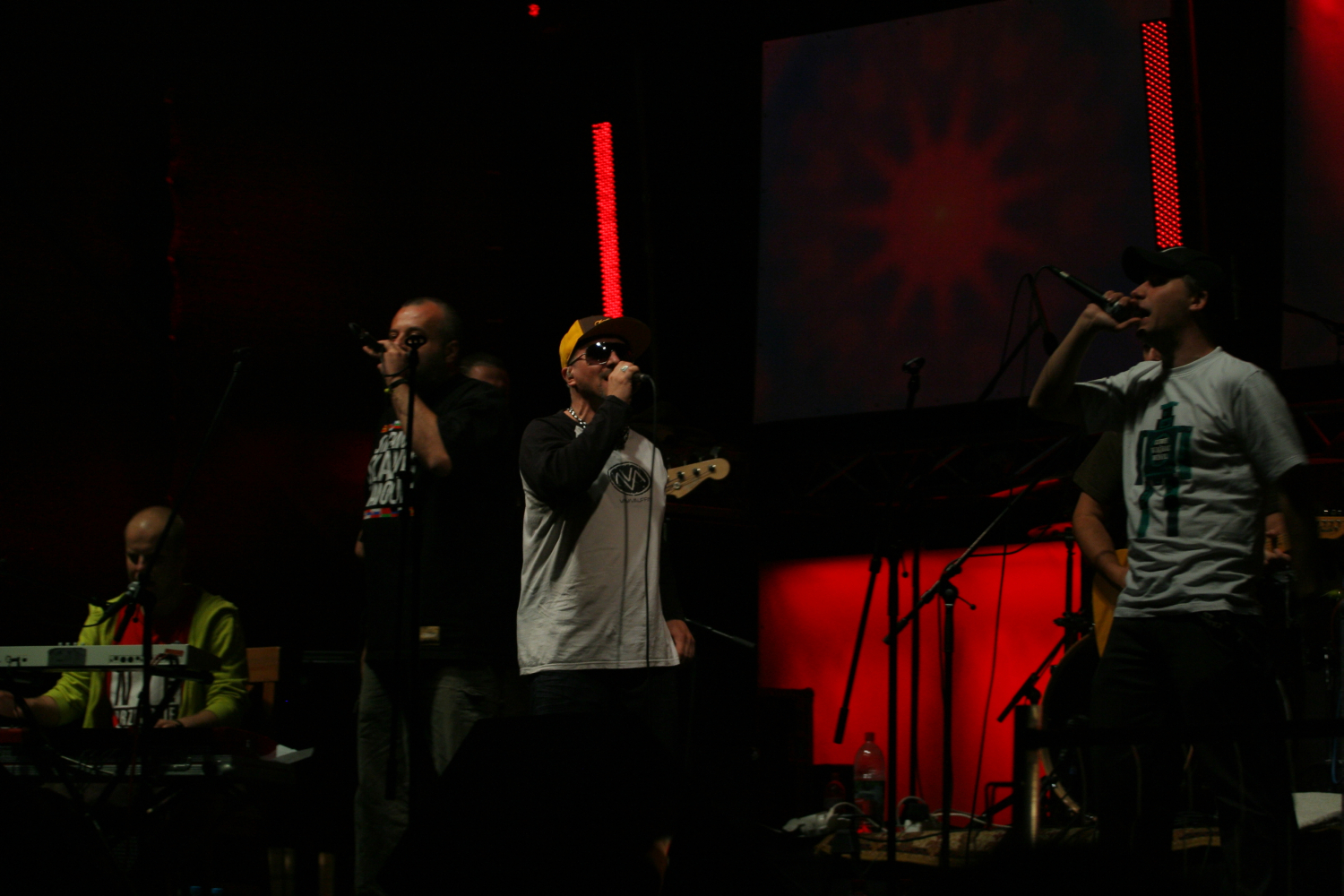A band performs on a dimly lit stage with red lighting. Four members of the band are holding microphones and singing. One sits at a keyboard on the far left, wearing a Laba 2011 graphic t-shirt. They are all dressed in casual clothes, including hats. Musical instruments and equipment are visible on the stage.