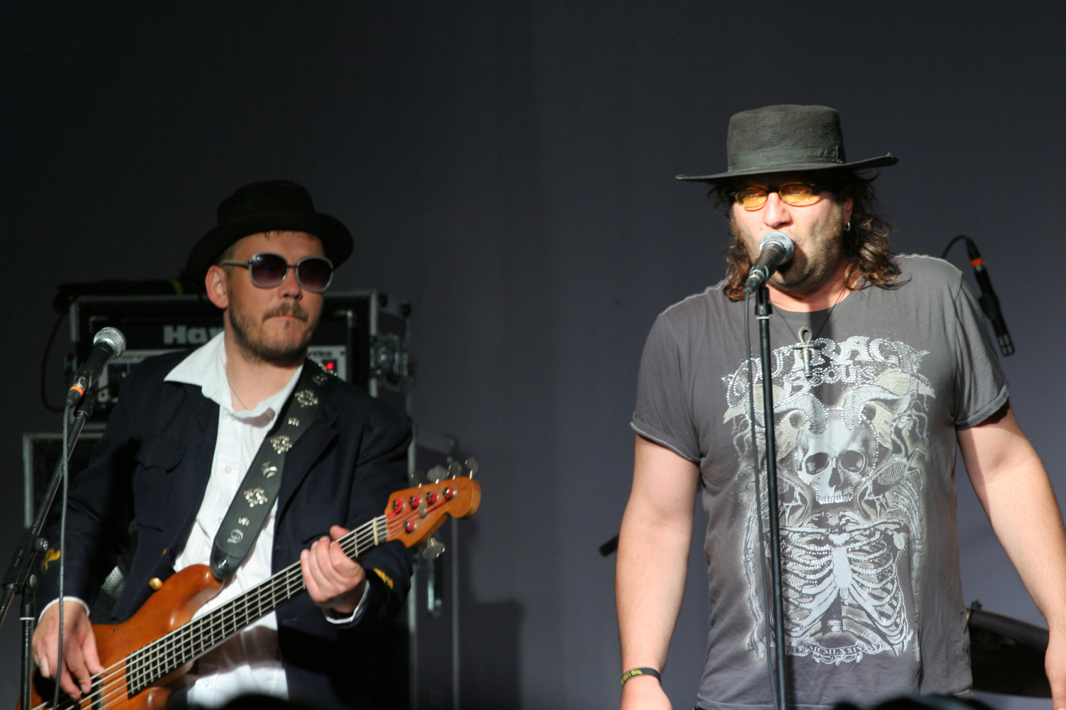 Two musicians performing on stage. The left musician, wearing a black hat, sunglasses, a white shirt, and a dark jacket, plays a bass guitar. The right musician, sporting a black hat and yellow-tinted sunglasses with a gray t-shirt featuring a skeleton design from Laba Daba 2011, sings into the microphone. Equipment is visible in the background.