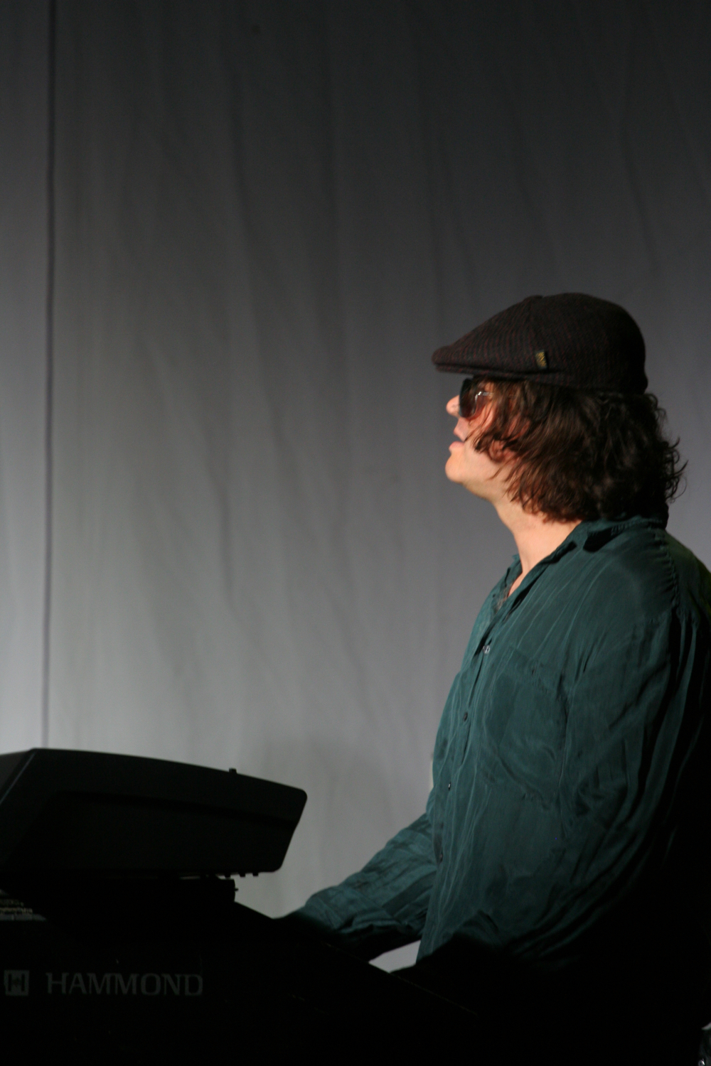 A person with shoulder-length curly hair, wearing a dark hat and sunglasses, is playing a Hammond keyboard. Dressed in a dark green, long-sleeve shirt and facing to the right against a plain, light-colored background, they evoke the vibrant spirit of Laba daba 2011.