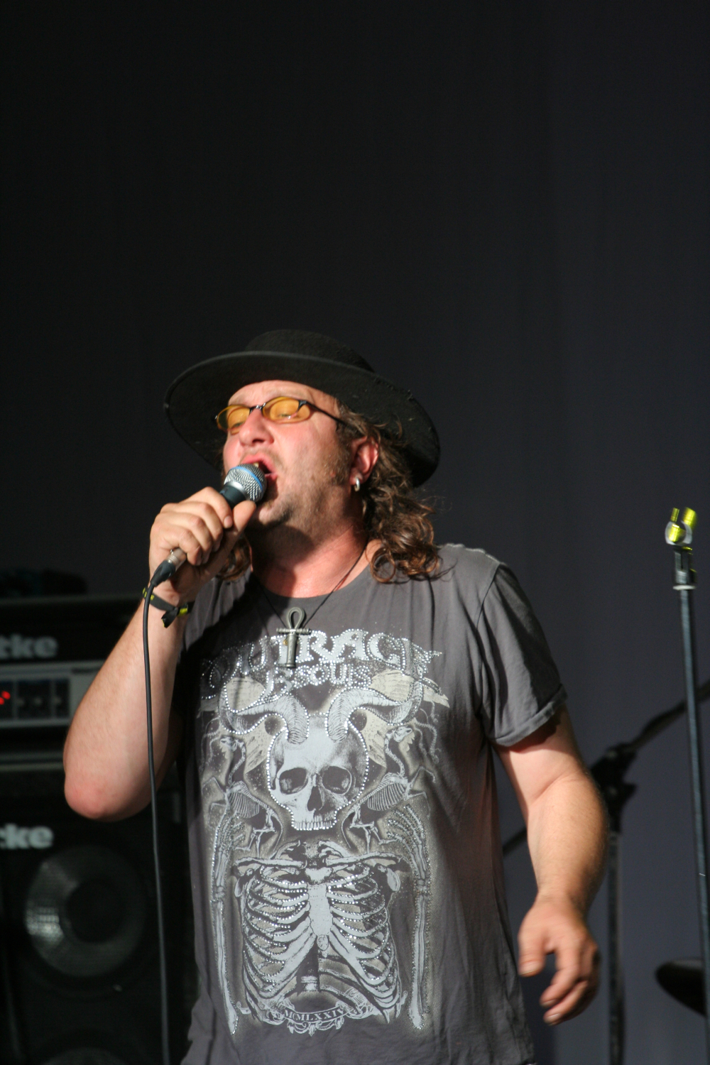 A man wearing a black hat, yellow-tinted glasses, and a t-shirt with a skeleton design reading "Outrage" is passionately singing into a microphone. His long hair flows past his black cross necklace. Behind him, audio equipment including a Roland speaker hints at the raw energy of this 2011 Laba daba scene.