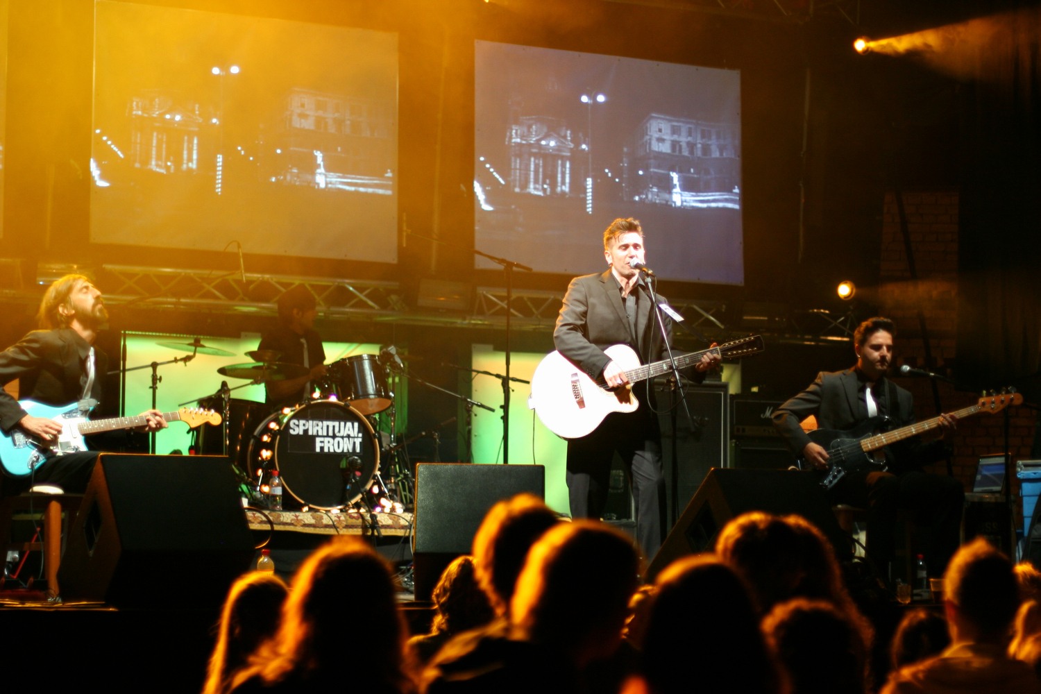 A band performs on stage under bright lights during the Laba Daba 2011 festival. The lead singer, playing a guitar, stands center stage in front of a microphone. On his left is a bassist, and on his right is a drummer with a drum set reading "Spiritual Front." The background features screens displaying black-and-white visuals.