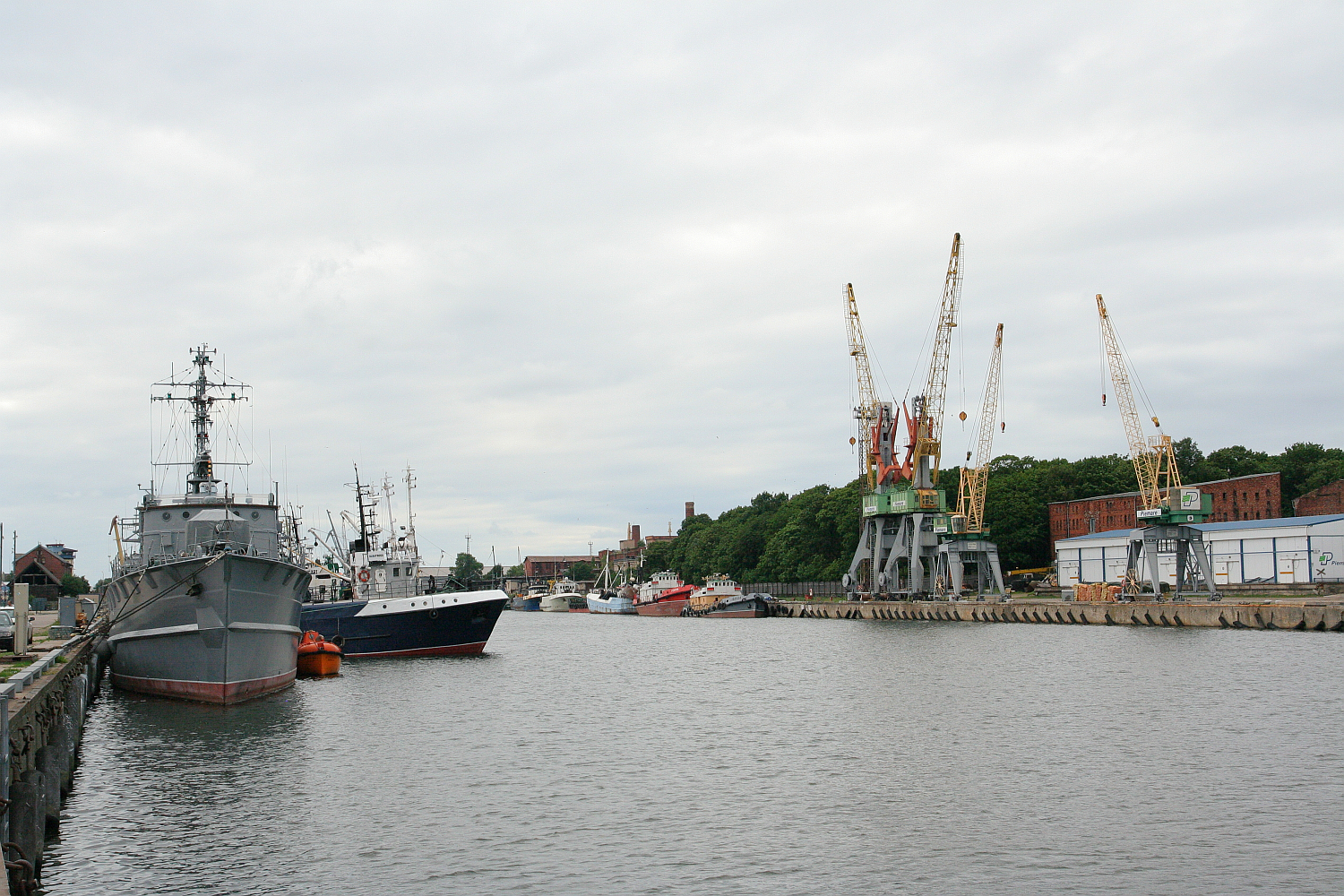 Naval and civilian vessels docked in a tranquil Liepāja harbor, flanked by towering cranes against a backdrop of overcast skies, hinting at bustling maritime industry and seafaring