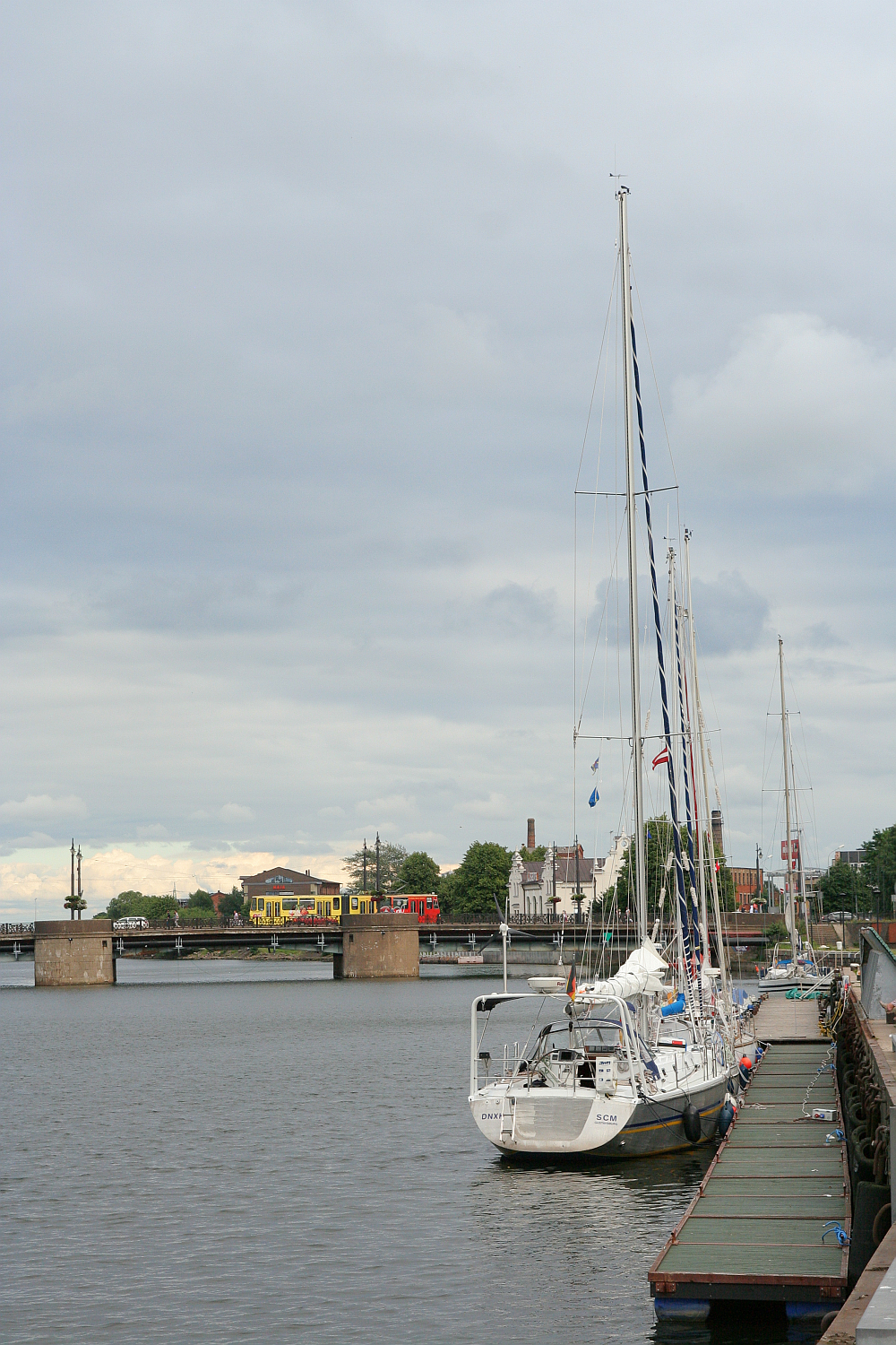A row of sailboats moored at a dock in Liepāja on a cloudy day, with calm water reflecting a bridge and buildings in the distance.
