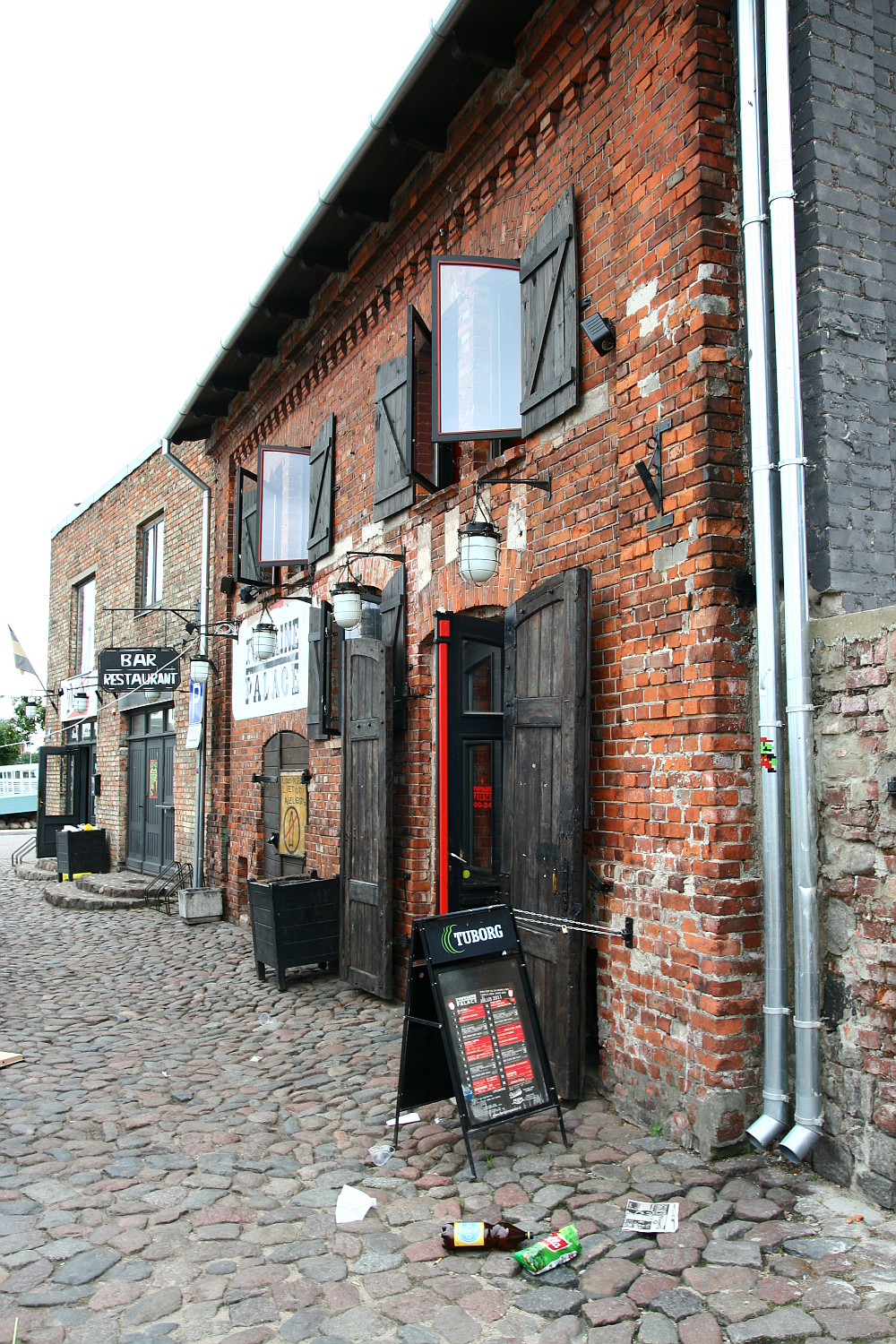 A rustic red brick building with wooden doors, hosting a bar and restaurant in Liepāja, on a cobblestone street; a blackboard menu and empty crates sit out front under clear skies