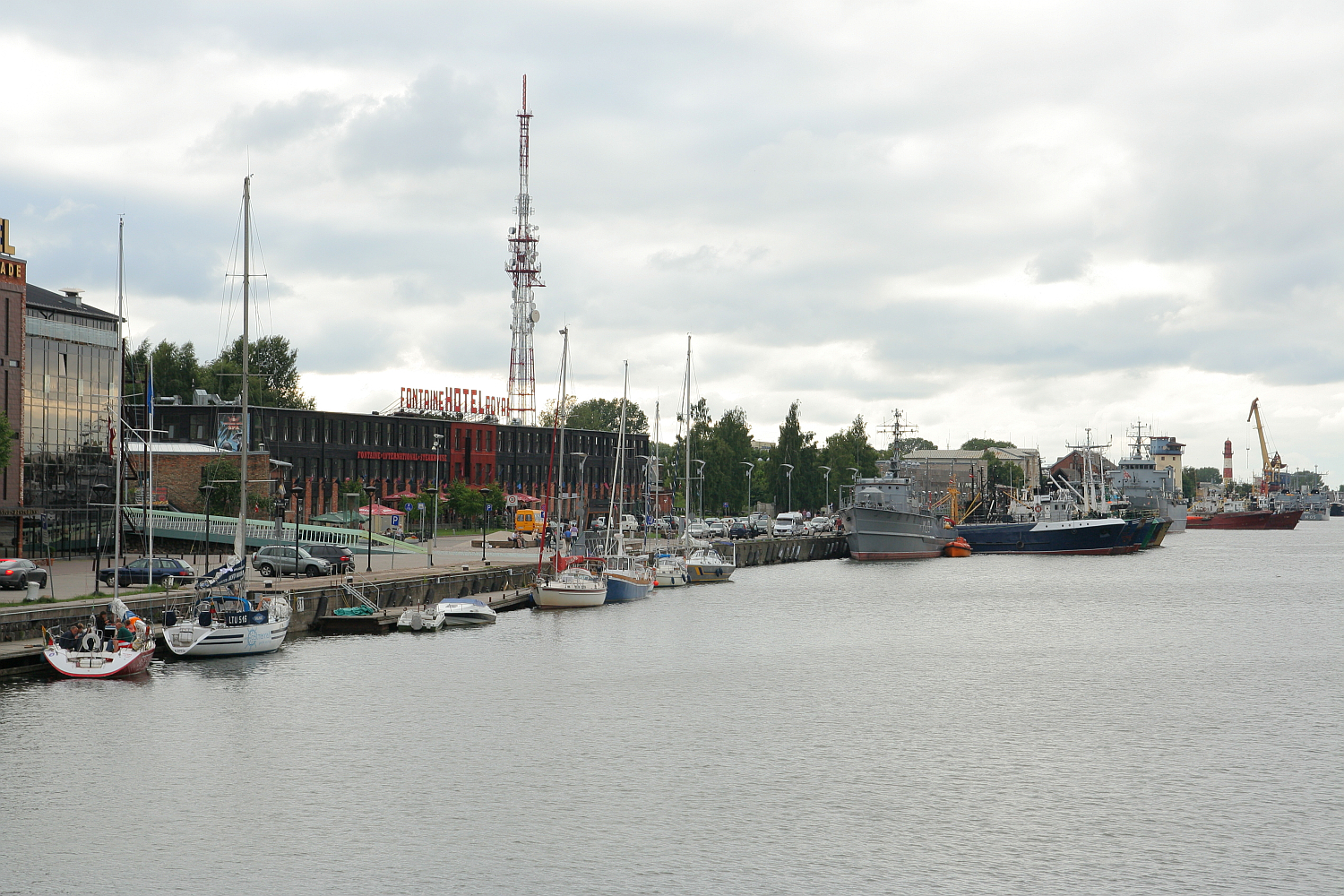 Cloudy skies over a bustling harbor in Liepāja, with an array of boats and ships moored along the quay; waterfront buildings and a towering communication mast stand in the background, suggesting