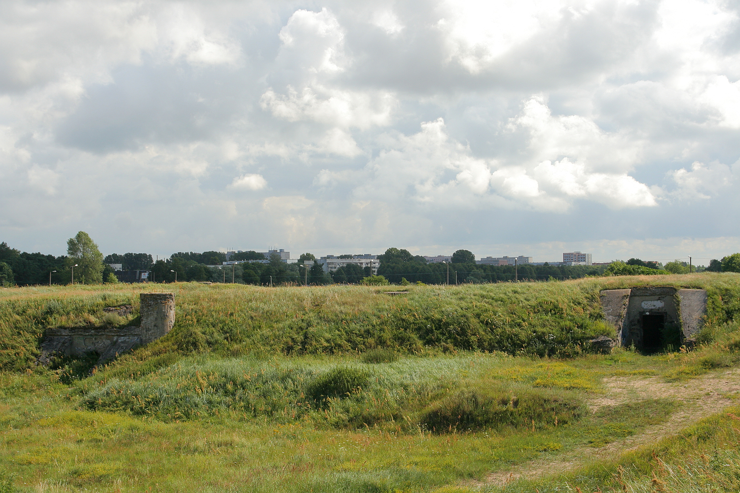 Verdant fields under a partly cloudy sky with remnants of old military bunkers near Liepāja, foregrounding a modern city skyline, illustrating a contrast between history, nature, and urban development