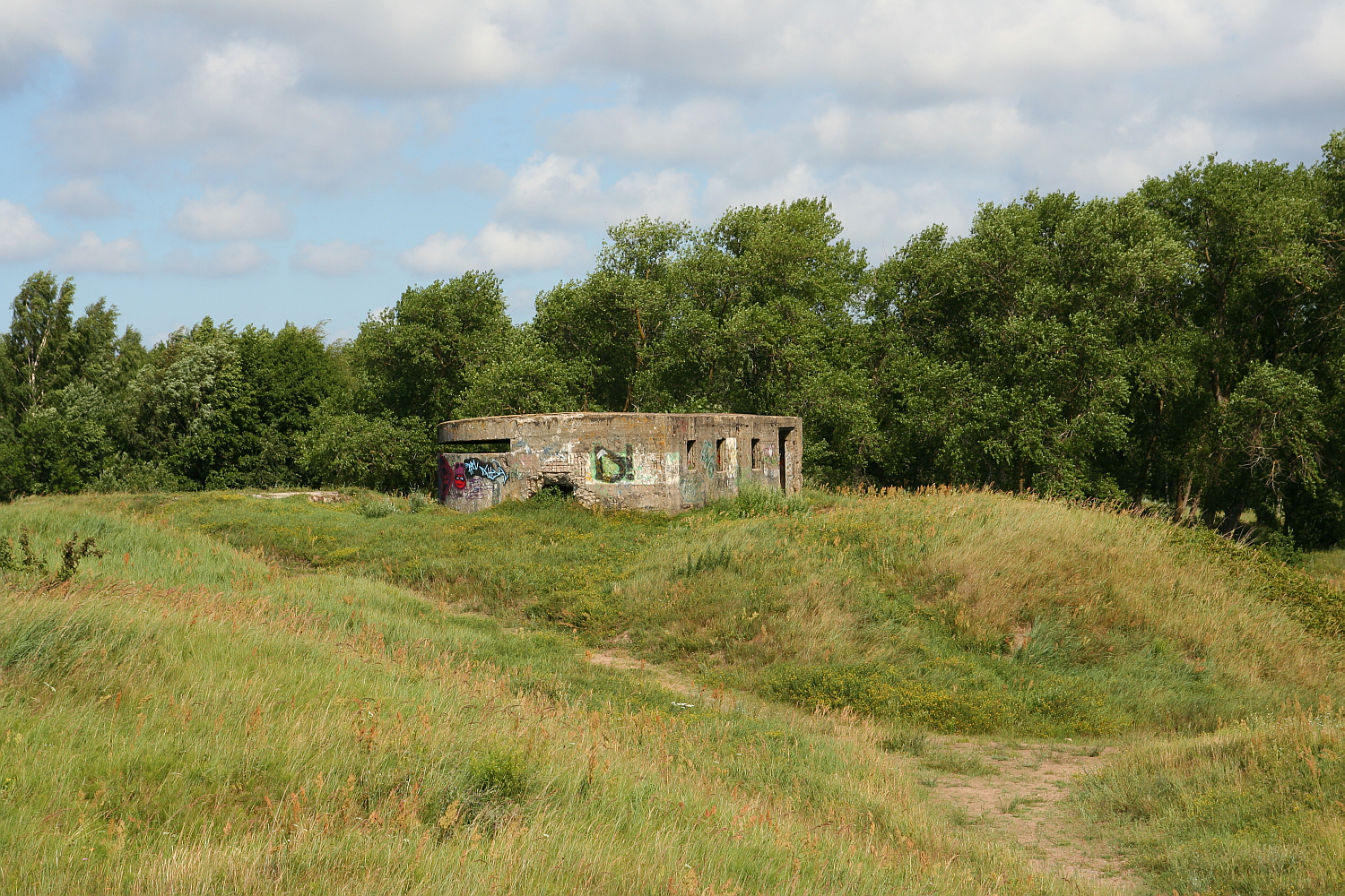 A weathered, graffiti-covered concrete building sits abandoned amidst verdant, grassy mounds on a sunny day with scattered clouds. This secluded structure in Liepāja, Latvia is surrounded by tall, lush green trees, and a narrow dirt path winds through the grass towards it, enhancing the scene's secluded feel.