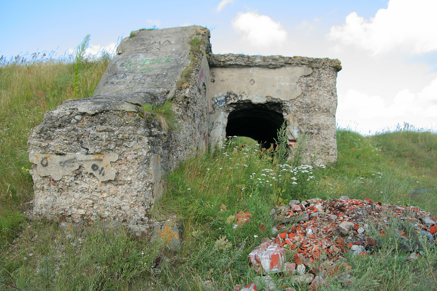 An abandoned concrete bunker in Liepāja, partially covered with graffiti, sits in a field with overgrown vegetation and a pile of rubble in the foreground under a partly cloudy sky.