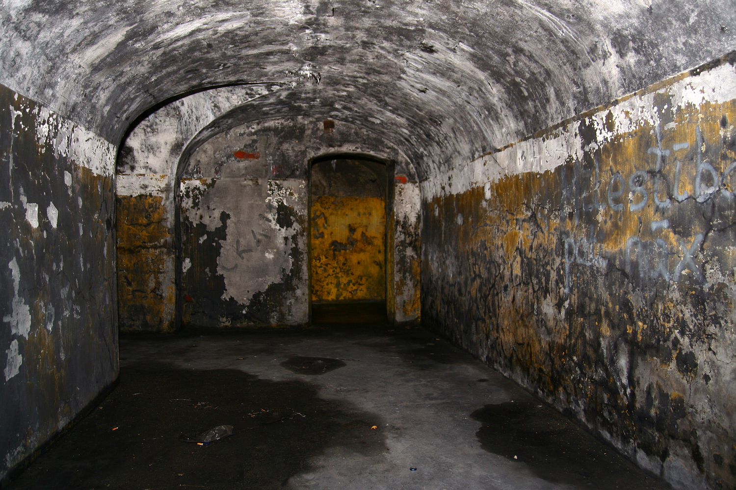 A dimly lit, abandoned curved tunnel in Liepāja with peeling, graffiti-marked walls leads to a weathered yellow door, creating an atmosphere of neglect and mystery.