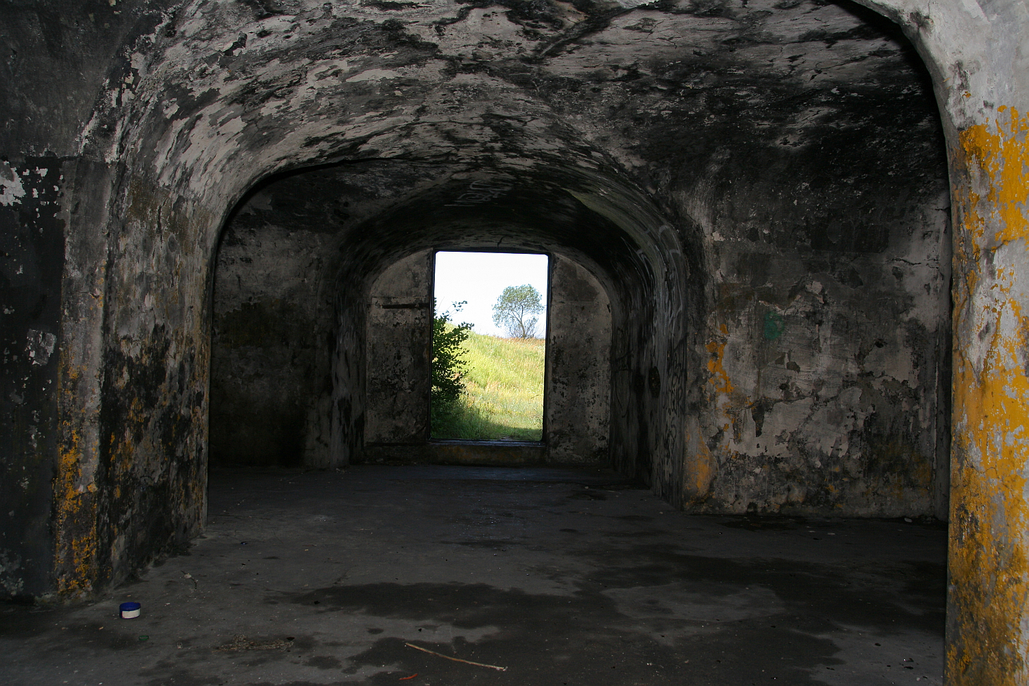 An old, arched stone tunnel with chipped paint and visible wear in Liepāja leads towards an outdoor exit, revealing a glimpse of greenery and a clear sky.