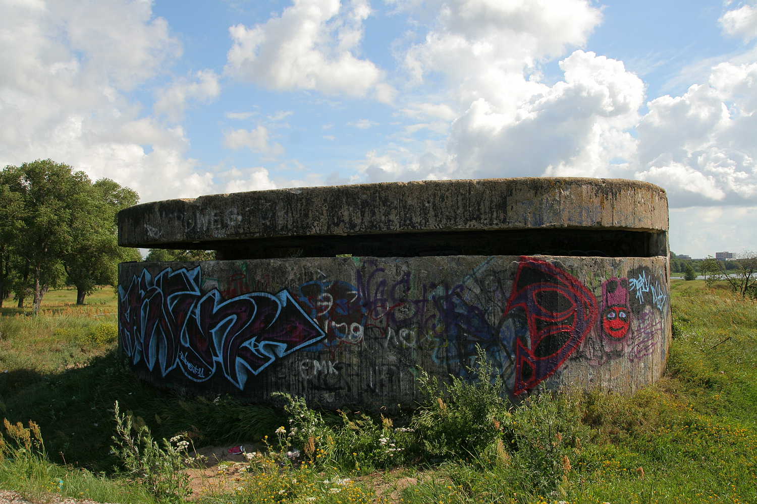 A weathered World War II concrete bunker covered in colorful graffiti, sitting under a cloudy sky in a grassy field near Liepāja, symbolizing a contrast between historical conflict and modern expression.