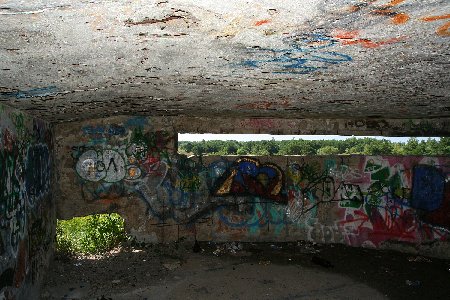 Graffiti-covered walls under a concrete structure in Liepāja, with colorful tags and drawings, offer a glimpse into local urban art and expression, contrasted by the bright, natural daylight peek