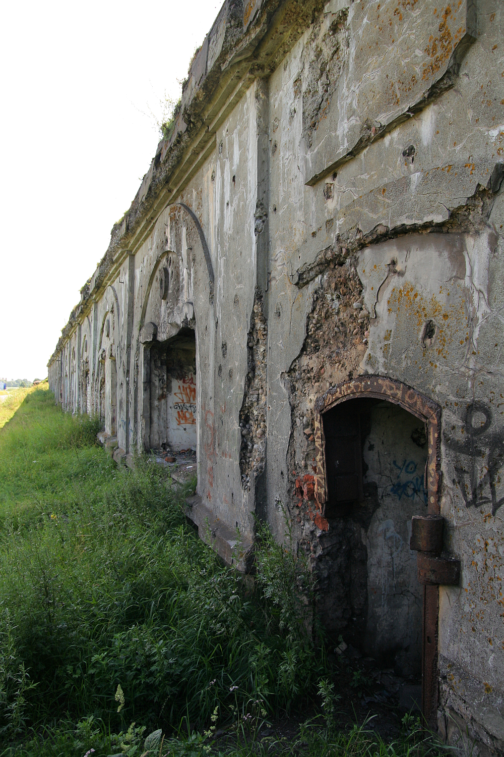 A long, weathered stone wall in Liepāja with large, arched openings in various stages of decay. The wall is covered in cracks and patches of missing plaster. Graffiti is visible on some sections. Overgrown grass and wild plants flank the base of the wall, leading to an open grassy area in the background.