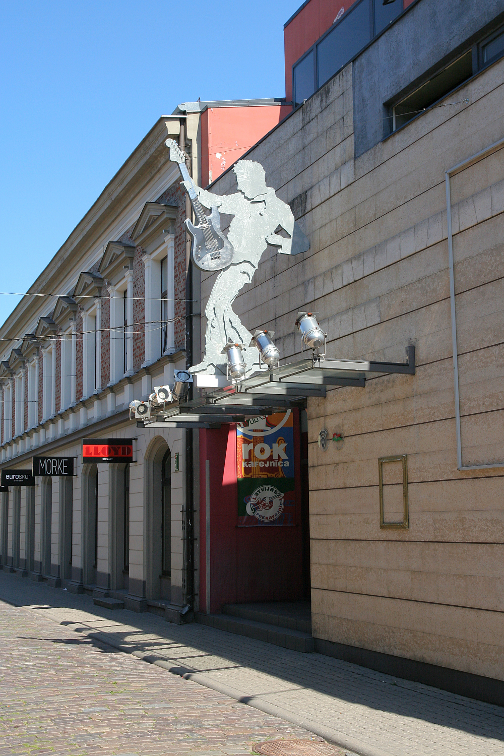 A dynamic metal sculpture of a guitarist extending from the facade of a building above a pedestrian street in Liepāja, showcasing a fusion of art and urban architecture under a clear blue sky.