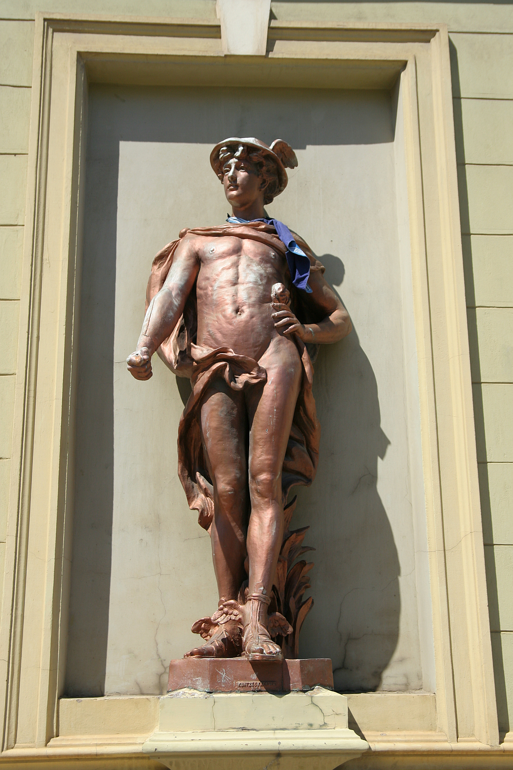 A bronze sculpture of a youthful, athletic male figure, assumedly an ancient hero or deity, is adorned with a draped cloth, set against a framed niche on a sunlit wall in Liepā