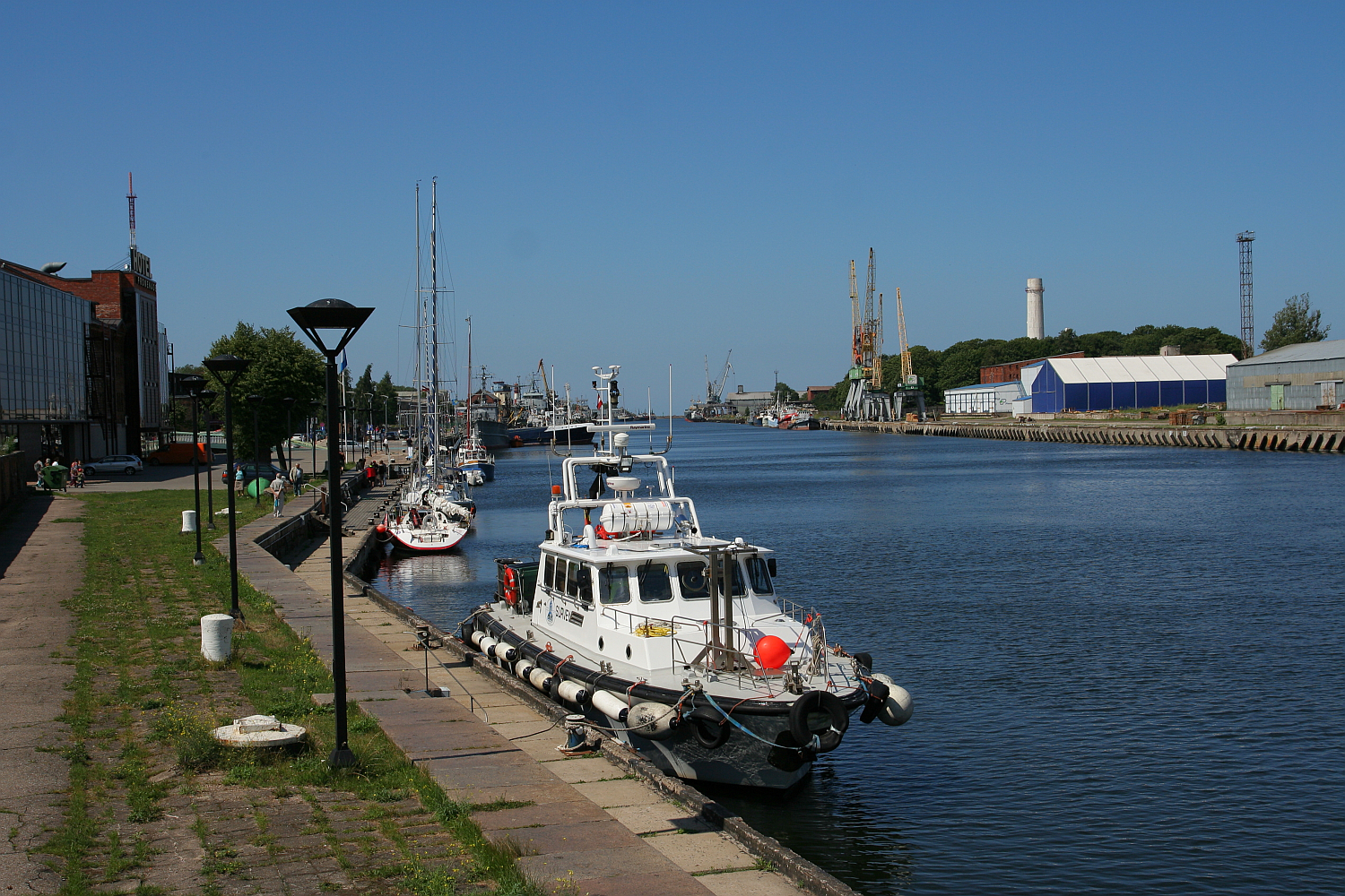 A sunny day at a calm port in Liepāja showing moored boats along a quay, with industrial buildings, cranes in the distance, and a clear blue sky overhead.