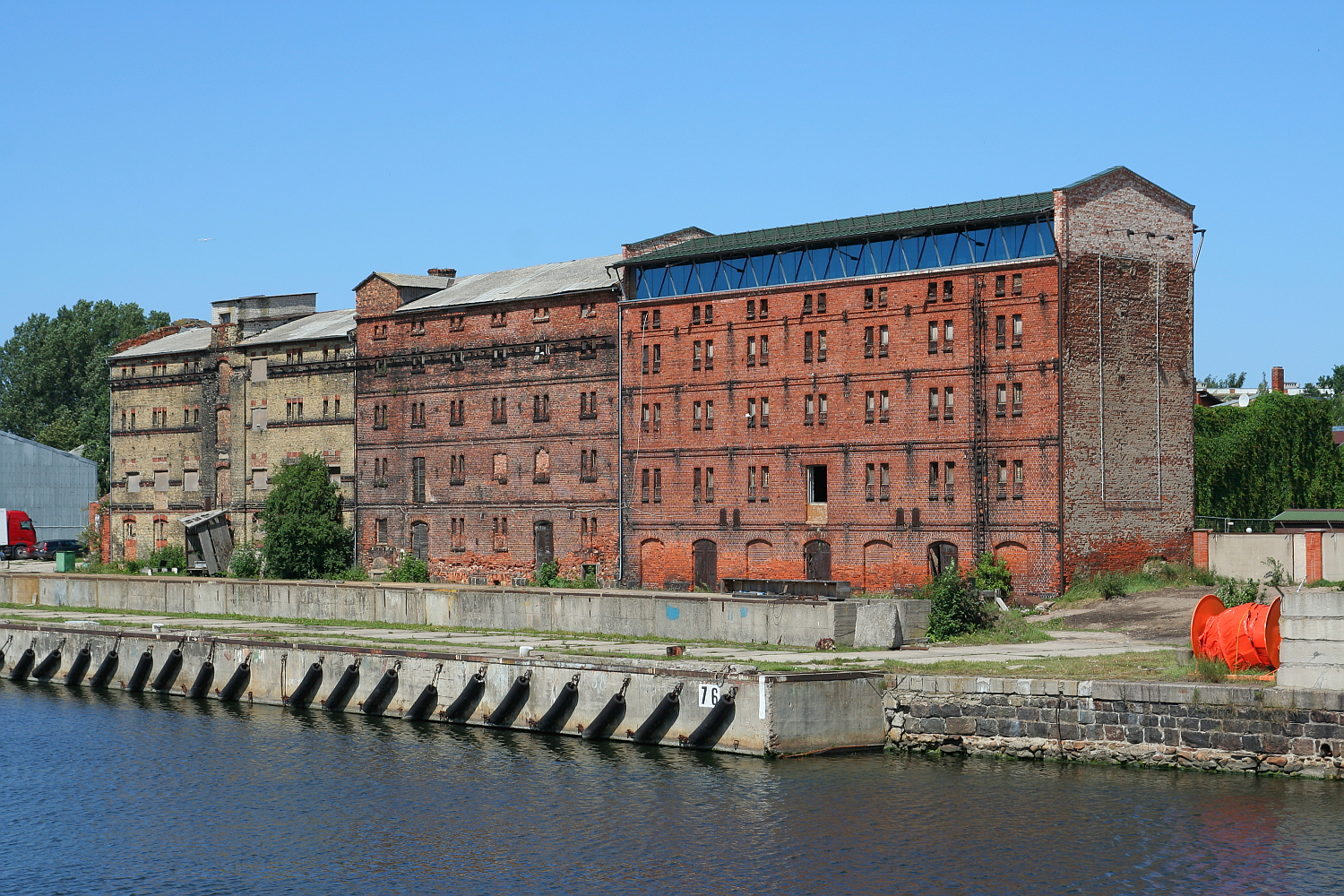 An imposing red brick industrial building, with signs of aging and disuse, stands by a calm waterway in Liepāja under a clear blue sky.