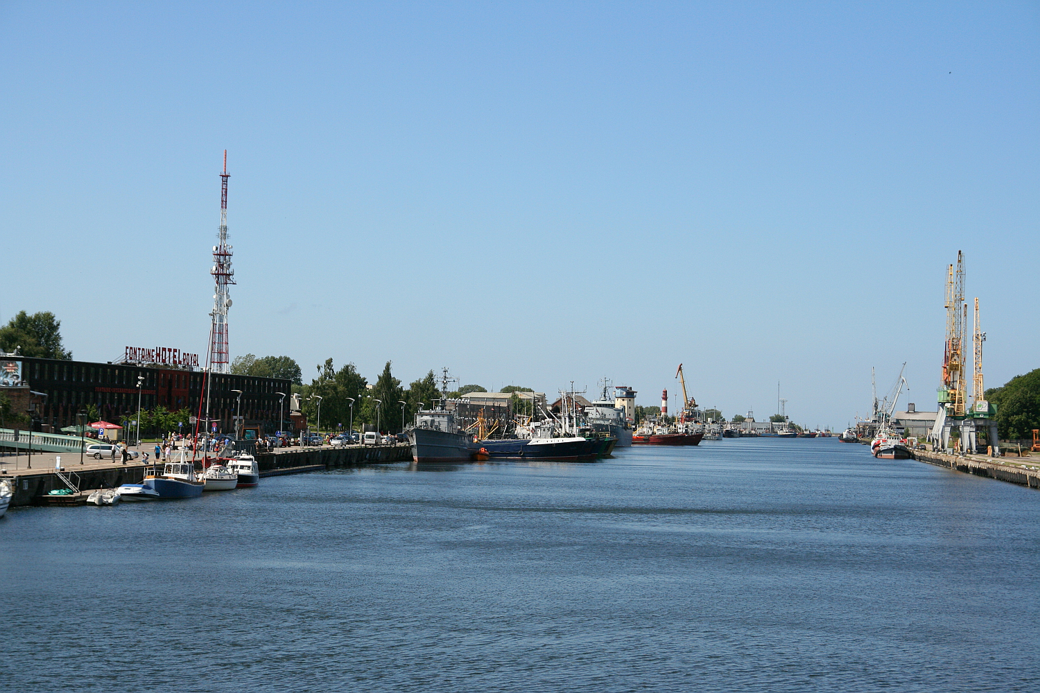 A clear day over the bustling port of Liepāja, with various ships docked along the quay and a blue sky reflecting in the calm water, while industrial cranes loom in the