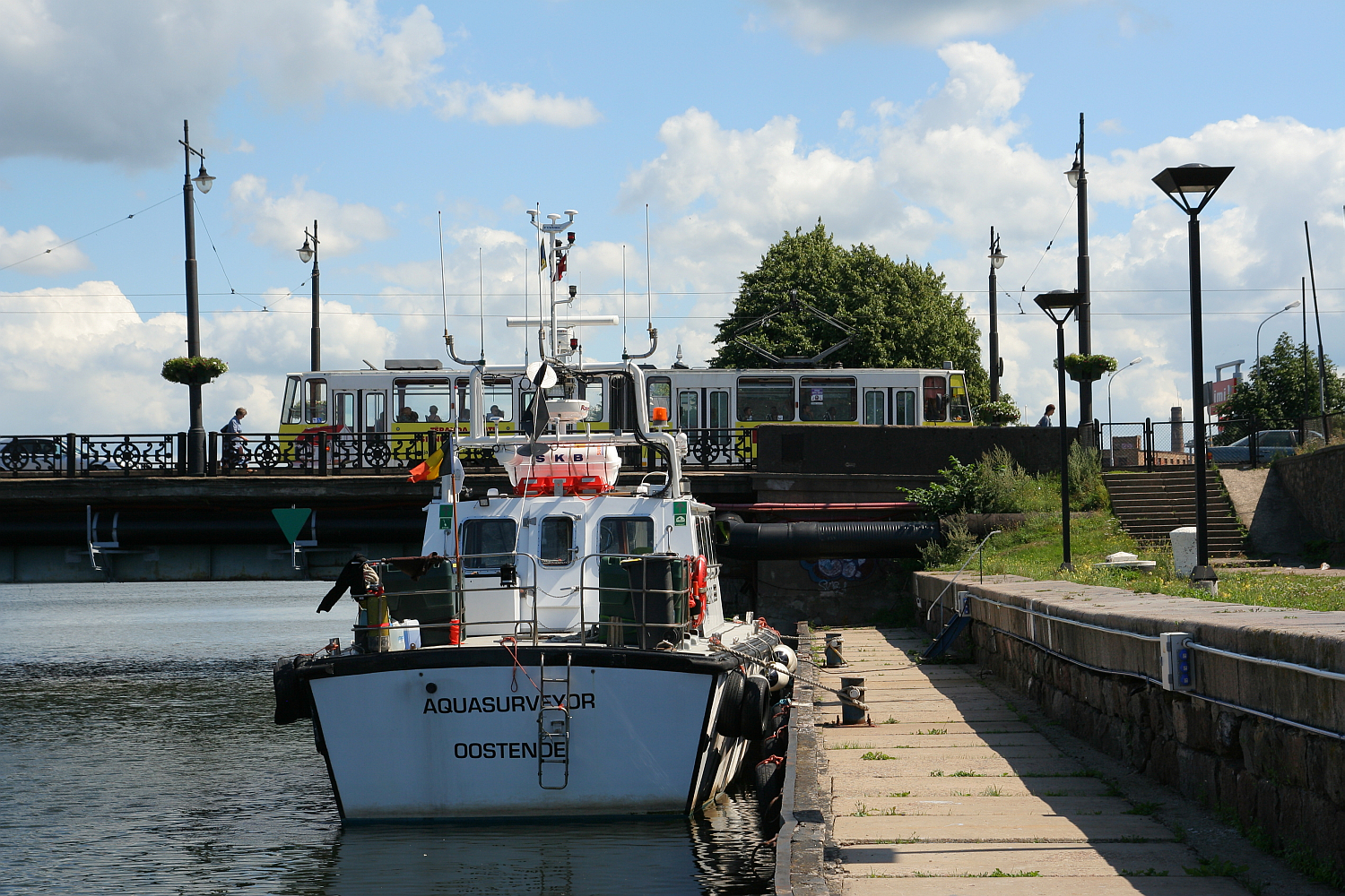 A survey vessel named "aquasurveyor" is moored on a calm riverbank in Liepāja, beneath a blue sky dotted with clouds, with a bridge and greenery in the