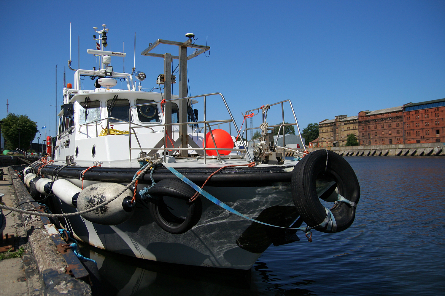 A tugboat with prominent fenders moored in a calm harbor in Liepāja, under clear blue skies, with urban buildings visible in the background.