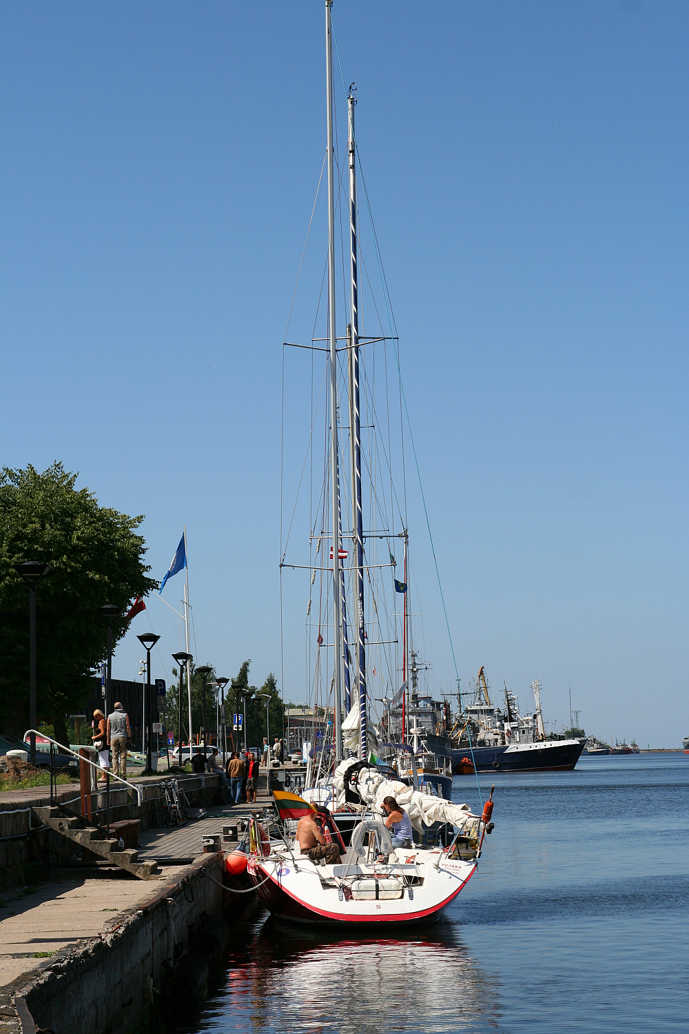 A sailboat with a red hull is docked at a waterfront promenade in Liepāja under clear blue skies, surrounded by calm waters and distant ships, inviting a day of maritime adventure.