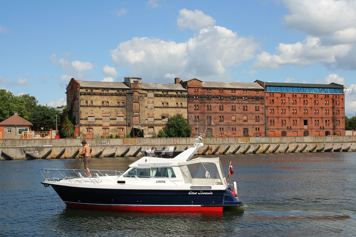 A luxury boat sails on a river in Liepāja, passing in front of historic brick warehouses under a blue sky dotted with clouds, illustrating a blend of recreational boating and industrial heritage.