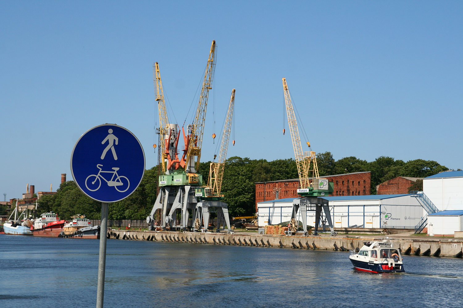 Sign indicating a pedestrian and bicycle lane in the foreground, with a waterfront and several cranes in the background. A small boat navigates the water near industrial buildings and trees under a clear blue sky, capturing a typical scene in Liepāja.