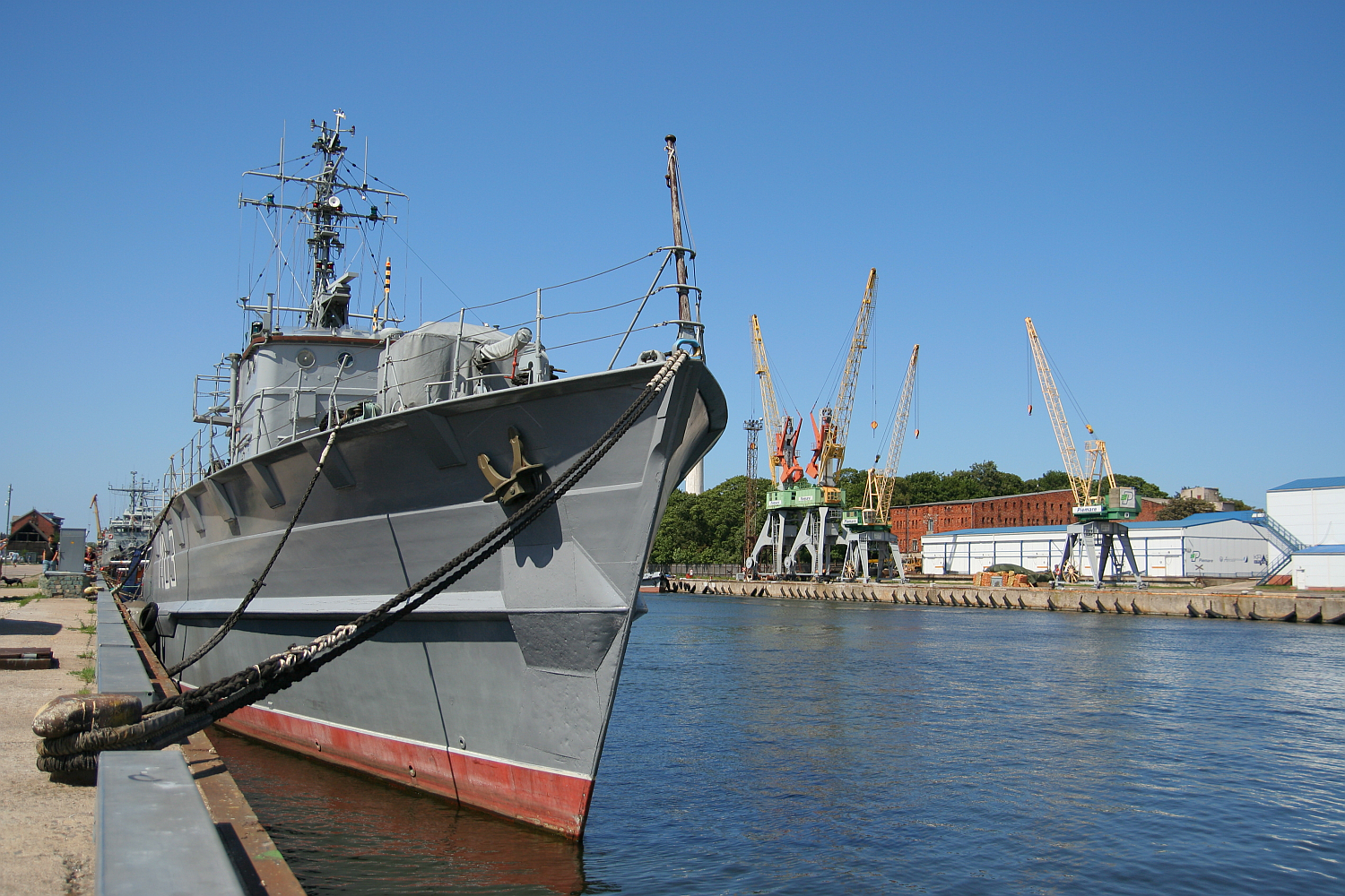 A military vessel moored at a calm dock in Liepāja, with towering cargo cranes in the background under a clear blue sky, signifying a blend of naval defense and commercial activity.
