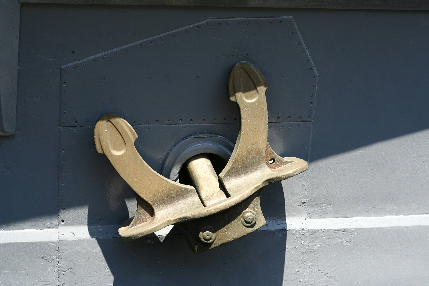 A shiny, gold-colored metal anchor is mounted on a dark grey surface with rivet details in Liepāja, suggesting a naval or maritime setting, with sunlight highlighting its contours.