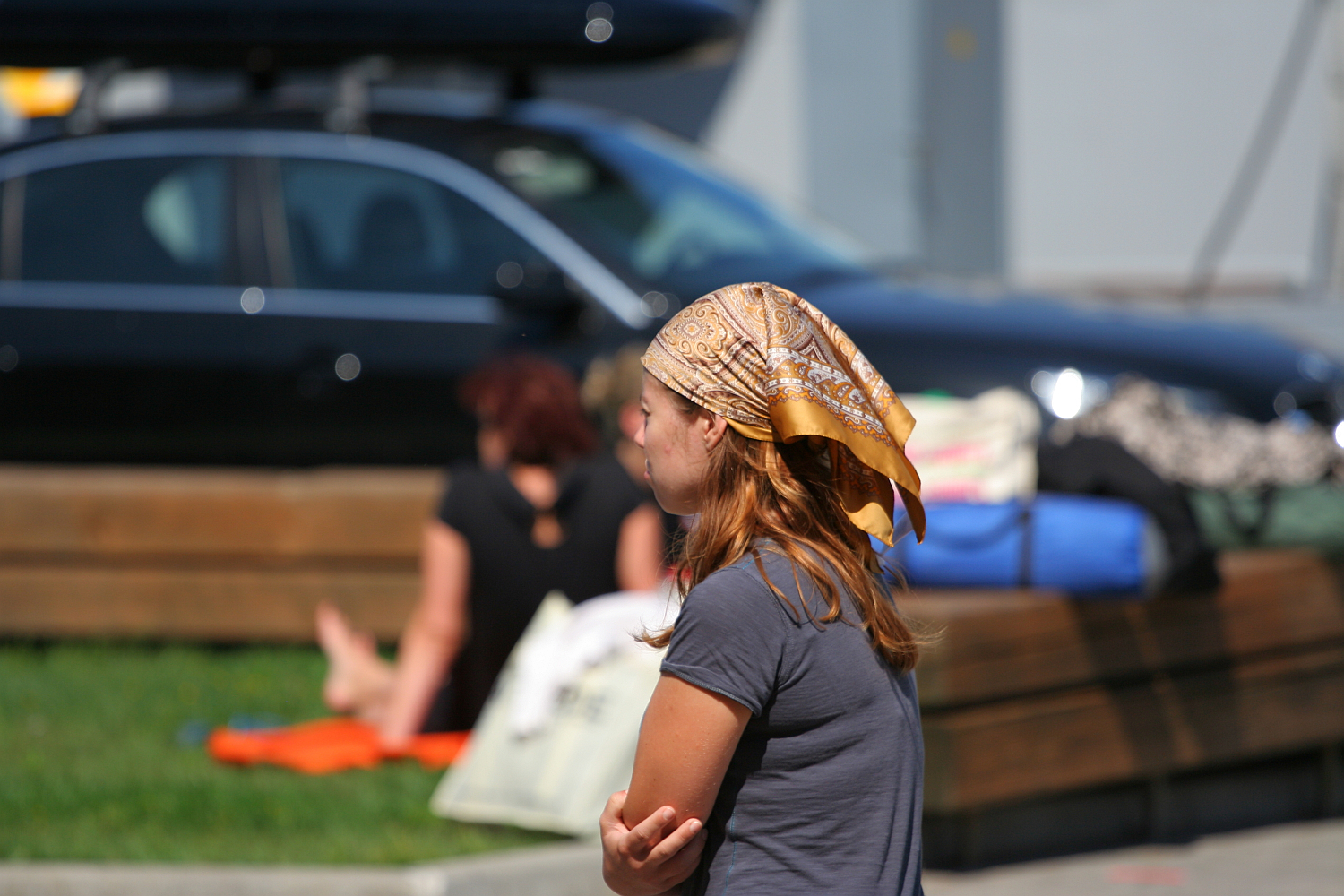 A person in a brown headscarf and grey top gestures with their hands while talking in Liepāja, with blurred figures sitting in the sunny background amidst parked cars.
