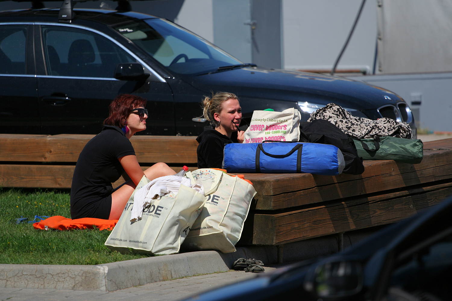 Two women sitting on the grass near a wooden ledge with several bags and a rolled-up sleeping bag in front of them. One woman with dark hair and sunglasses sits on an orange mat, while the other, with blonde hair, talks animatedly about their trip to Liepāja. A parked black car is visible in the background.