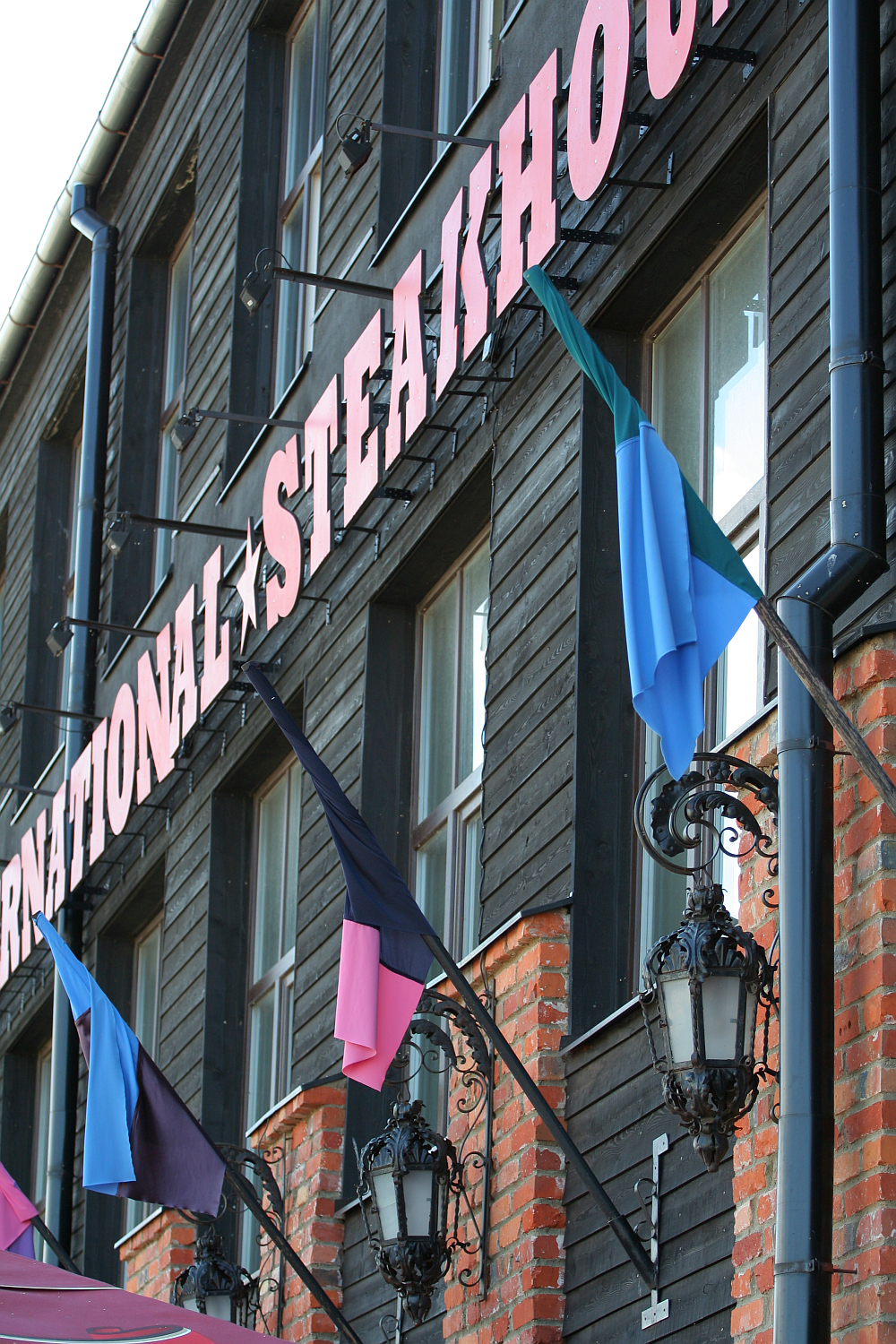 Facade of a steakhouse in Liepāja with bold signage, featuring black wood paneling and vibrant blue and pink flags under a clear sky, complemented by ornamental black street lamps.