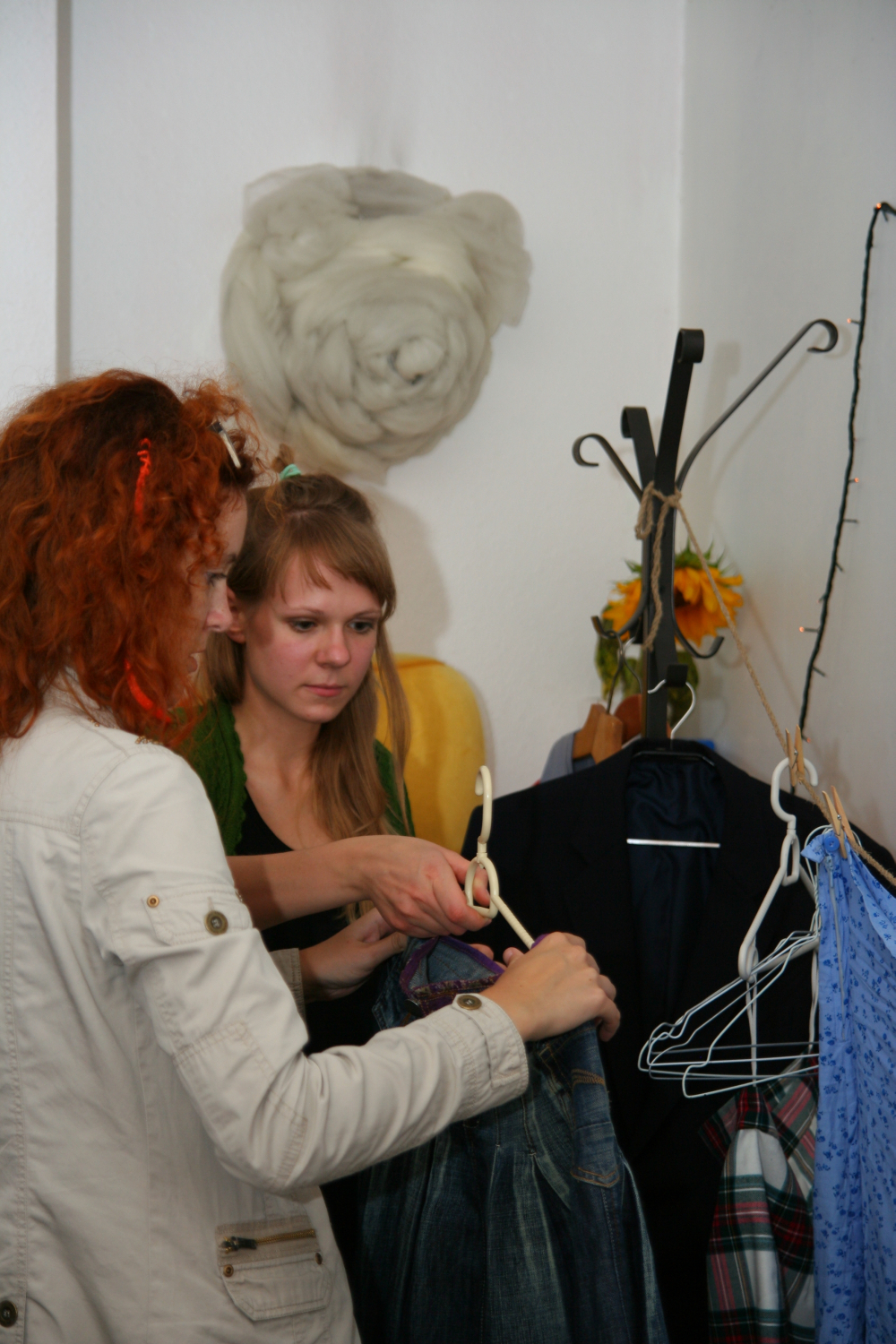 Two women examine clothing in a small, cluttered space. One woman, with curly red hair and glasses, holds a hanger with jeans. The other woman, with straight blonde hair and bangs, looks on. Behind them is a coat rack with hanging clothes, likely preparing for a Maiņas punkts preparty event.