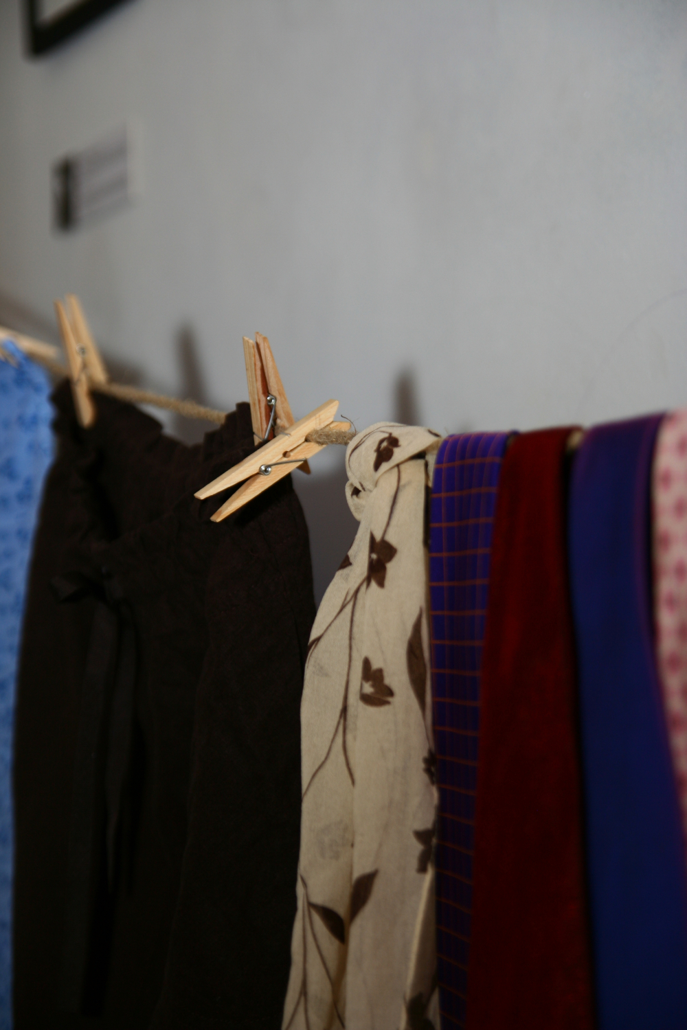 Several pieces of cloth, including a wooden clothespin-hung brown fabric, a floral-patterned beige scarf, and colorful garments in blue, red, and purple with a checked pattern, are drying on a clothesline indoors against a grey wall. The background includes a blurred framed Maiņas punkts sign.
