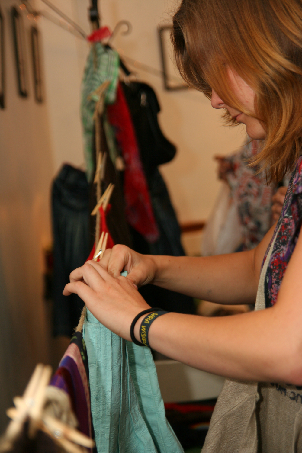 A woman with medium-length brown hair is looking closely at a green garment she is holding with both hands. She wears a black wristband and a colorful scarf. In the background, clothes on hangers and pegs are visible, hinting that this preparty shopping session might be happening at Maiņas punkts.