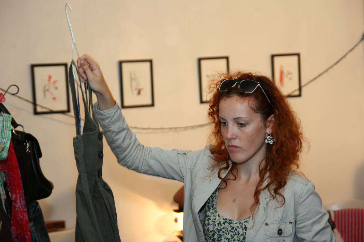 A person with curly red hair is holding up a piece of clothing on a hanger for inspection, perhaps picking out their preparty outfit. They are wearing a light gray jacket, sunglasses on their head, and earrings. Behind them at this Maiņas punkts, there are framed pictures on the wall and some clothes hanging on a rack to the left.