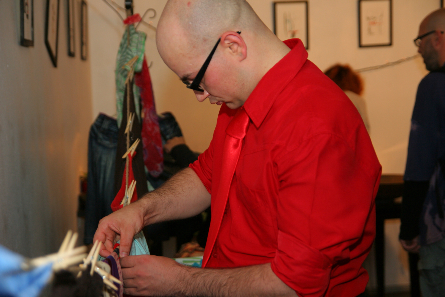 A bald man in a red shirt and tie, wearing black-rimmed glasses, is fixing or adjusting items clipped onto a line with colorful clothespins indoors. Framed pictures decorate the white wall behind him, adding an artistic flair to the preparty scene. Other people are present in the background.