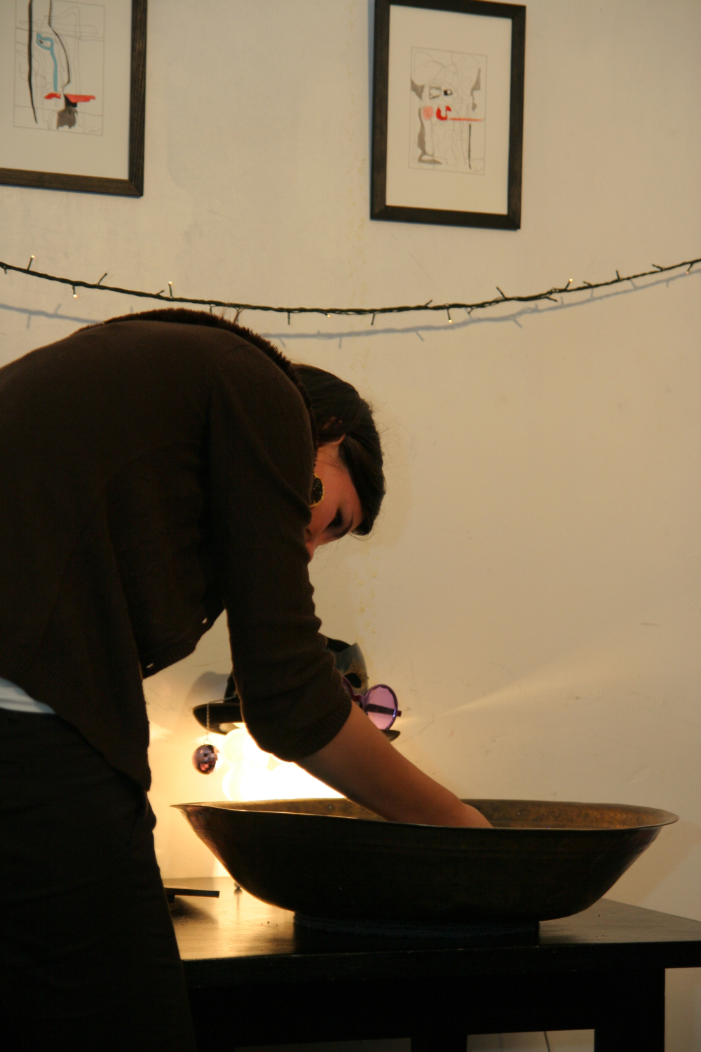 A person with dark hair, wearing a brown top, leans over a large, shallow basin on a dark table. They appear focused on something in the basin. Behind them, two framed abstract drawings are hung on a light-colored wall, and a string of lights decorates the space, setting the perfect preparty mood.