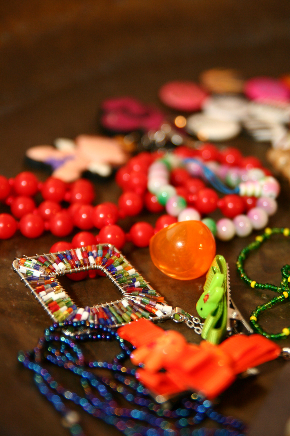 A close-up of assorted colorful jewelry pieces placed on a surface. In the foreground are beaded necklaces with red, green, and blue beads, a square pendant adorned with multicolored beads, and an orange, translucent bauble. Perfect for a preparty look, additional pieces blurred in the background include beads and various trinkets.