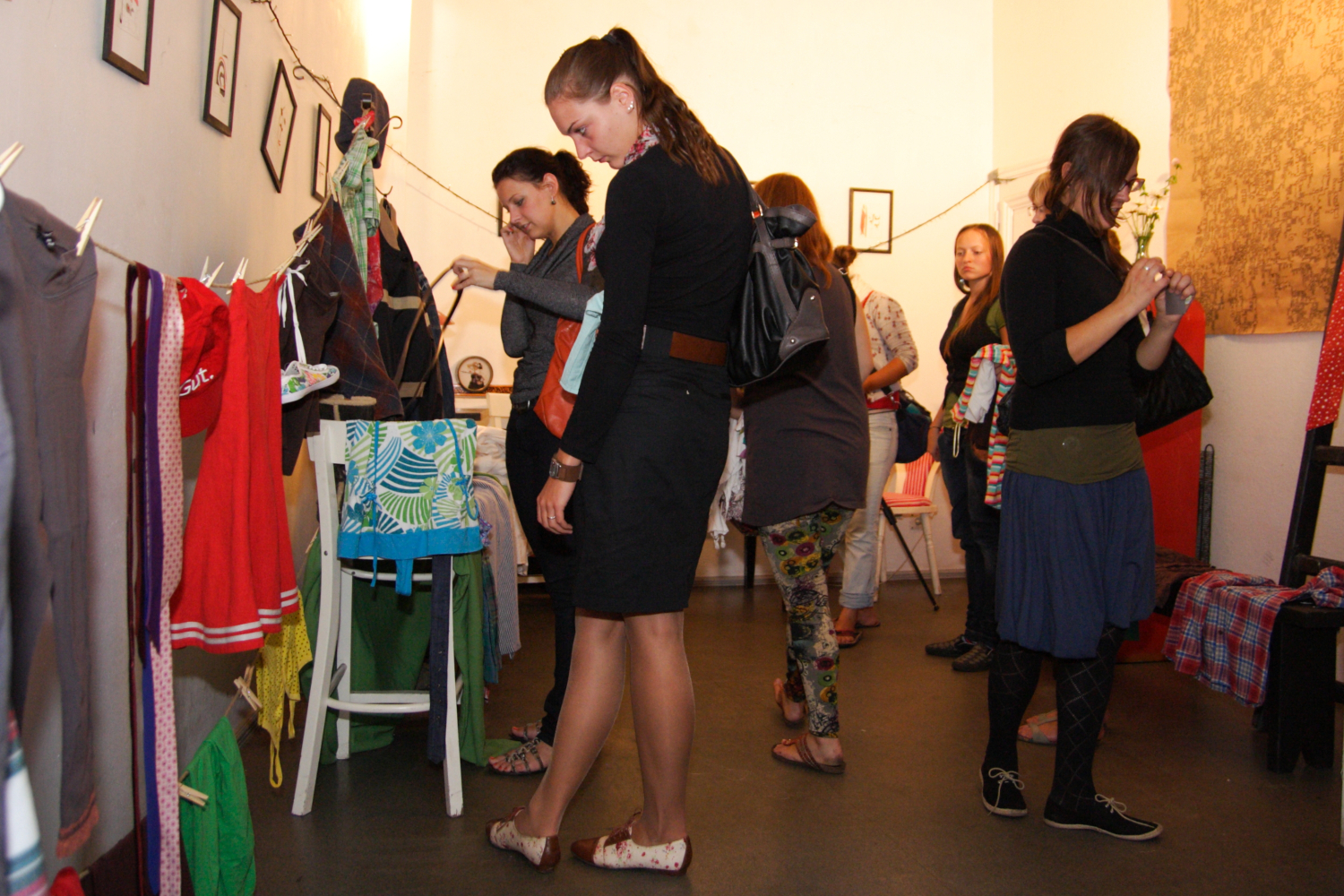 People browsing clothing and accessories in a small, indoor market or shop. The room is dimly lit with framed pictures on the walls. Clothes hang on a line and a rack. Individuals are engaged and examining items in this prepary Maiņas punkts. The atmosphere seems casual and friendly.
