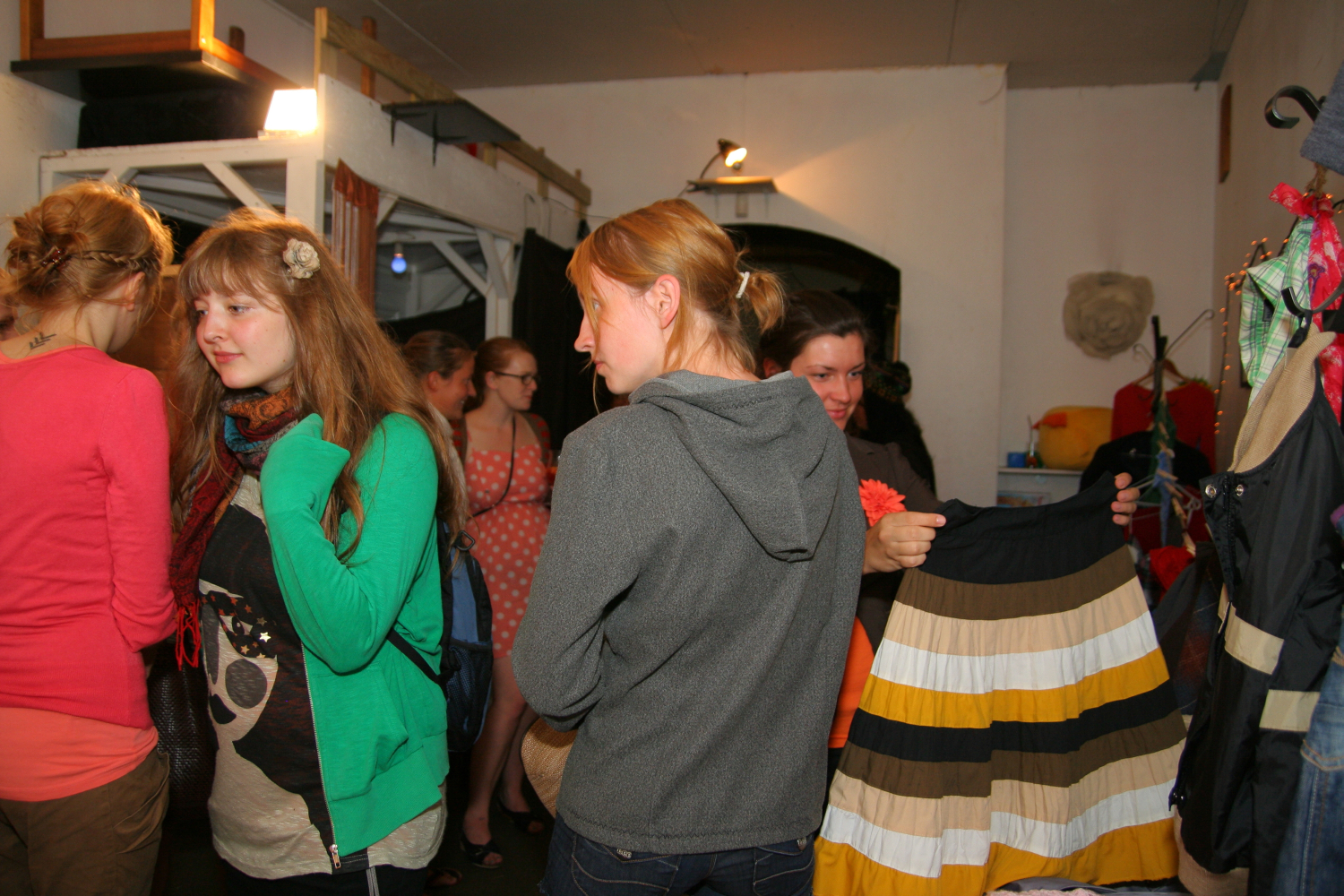 People shop at a crowded indoor market or Maiņas punkts. A person in a gray hoodie holds a striped skirt, while others browse nearby. Clothing hangs on racks, and various items are scattered throughout the space. The atmosphere appears lively and casual, as if preparing for a preparty.